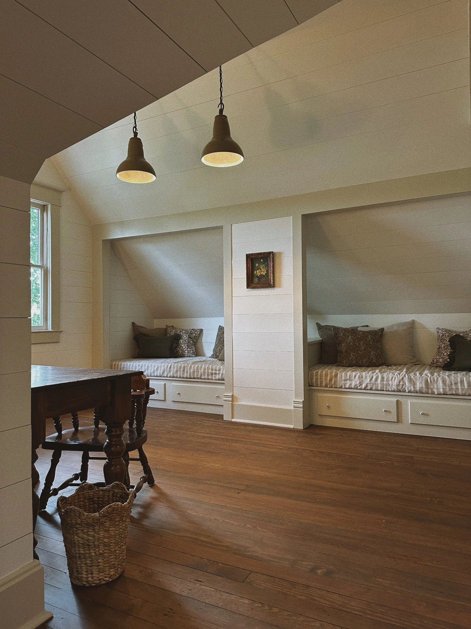 Interior of a room with white wood-paneled walls and ceilings, featuring two built-in beds with striped bedding and pillows, a small framed floral painting on the wall, wooden flooring, a window on the left letting in natural light, a dark wooden tab