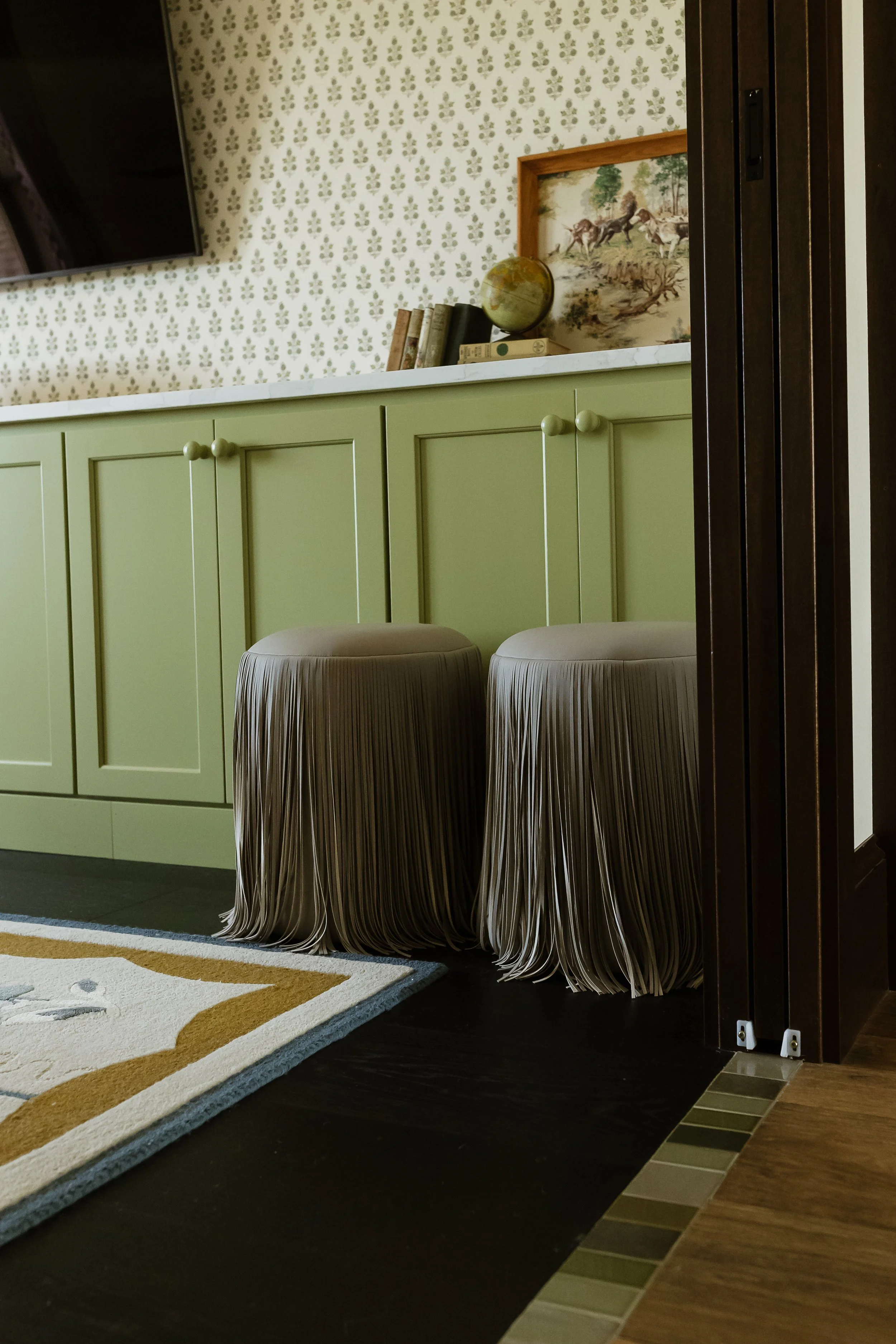 A room with a green cabinet, two beige fringe stools, a patterned rug, a wall with wallpaper, a wall-mounted TV, and a shelf with books, a framed picture of horses, and a globe.