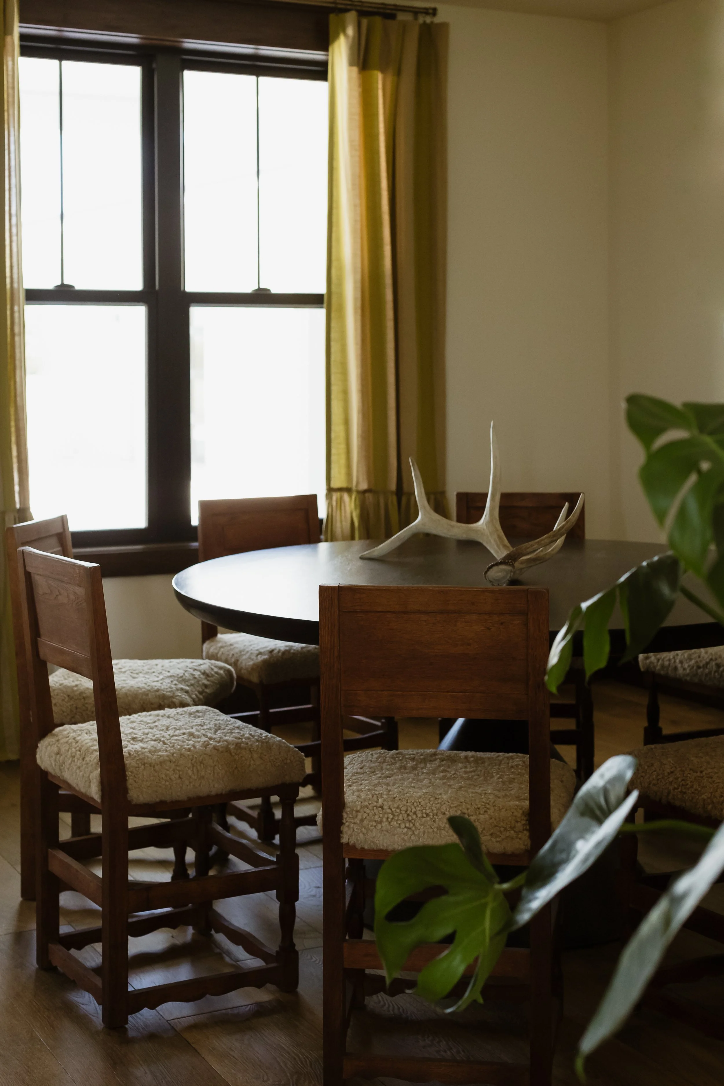 A dining room with a round wooden table and multiple wooden chairs with cushioned seats, a large window with yellow curtains, and a decorative antler on the table.