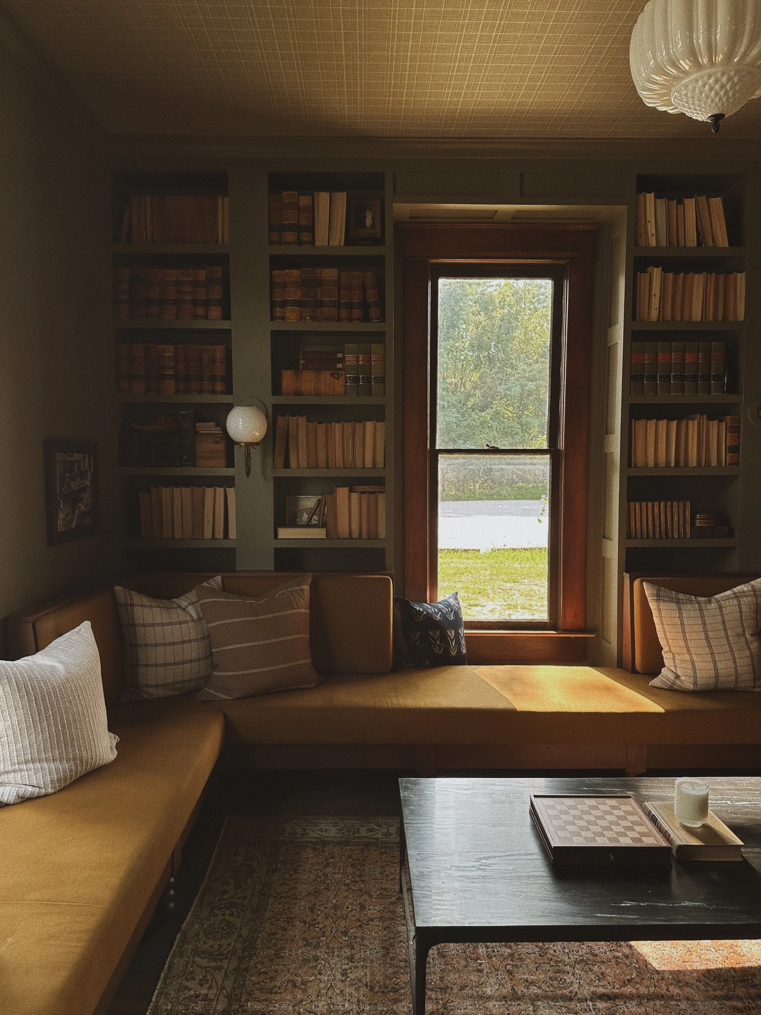A cozy living room with a large window showing outdoor greenery, built-in bookshelves filled with books, a beige sofa with plaid pillows, and a dark wooden coffee table with a chessboard and candles.