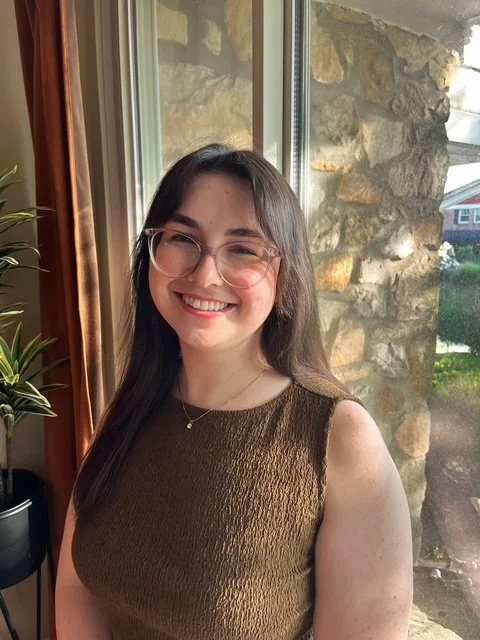 A young woman with long dark hair, glasses, and a brown sleeveless top smiling indoors near a window with a stone wall outside.