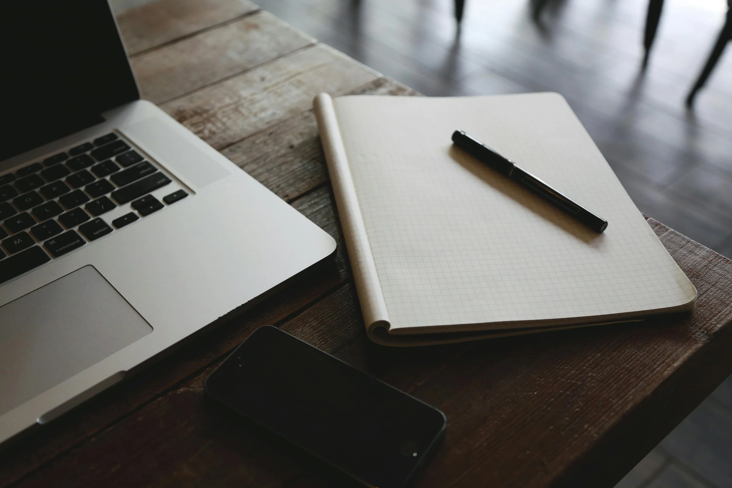 A wooden table with a silver MacBook, an open blank notebook with a black pen on top, and a black smartphone.