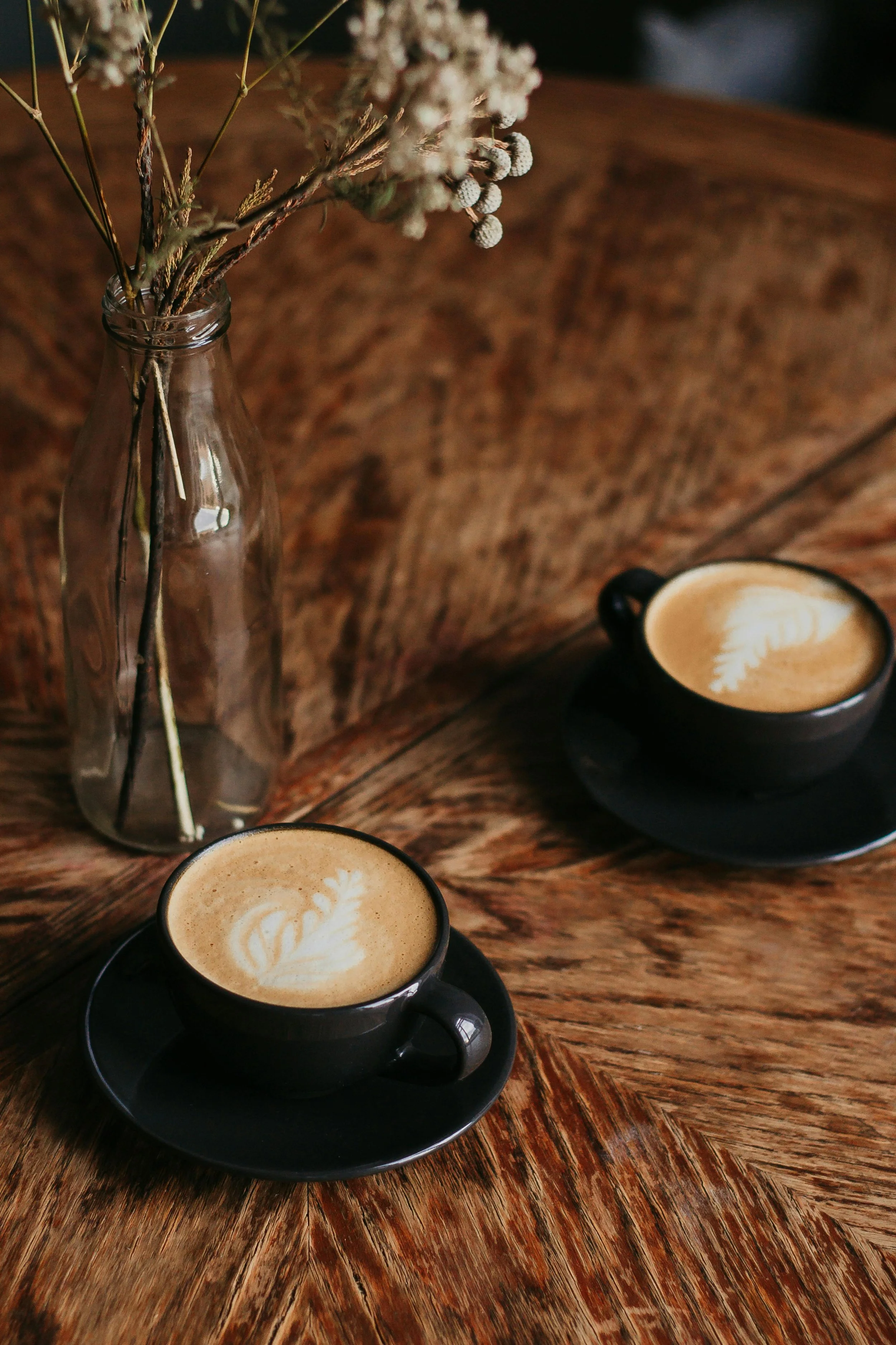A wooden table with two black cups of coffee with latte art and a clear glass bottle with dried flowers.
