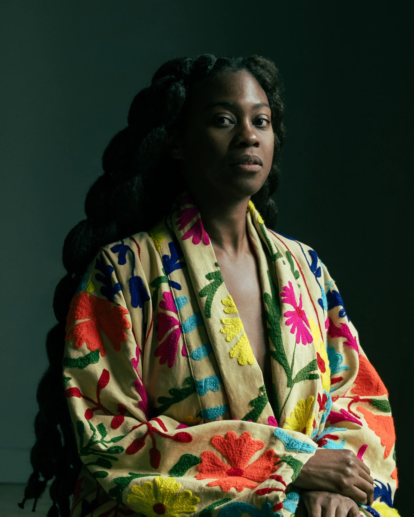 Portrait of a woman with dark hair styled in twists, wearing a colorful embroidered robe with floral patterns in red, blue, yellow, and green, set against a dark background.
