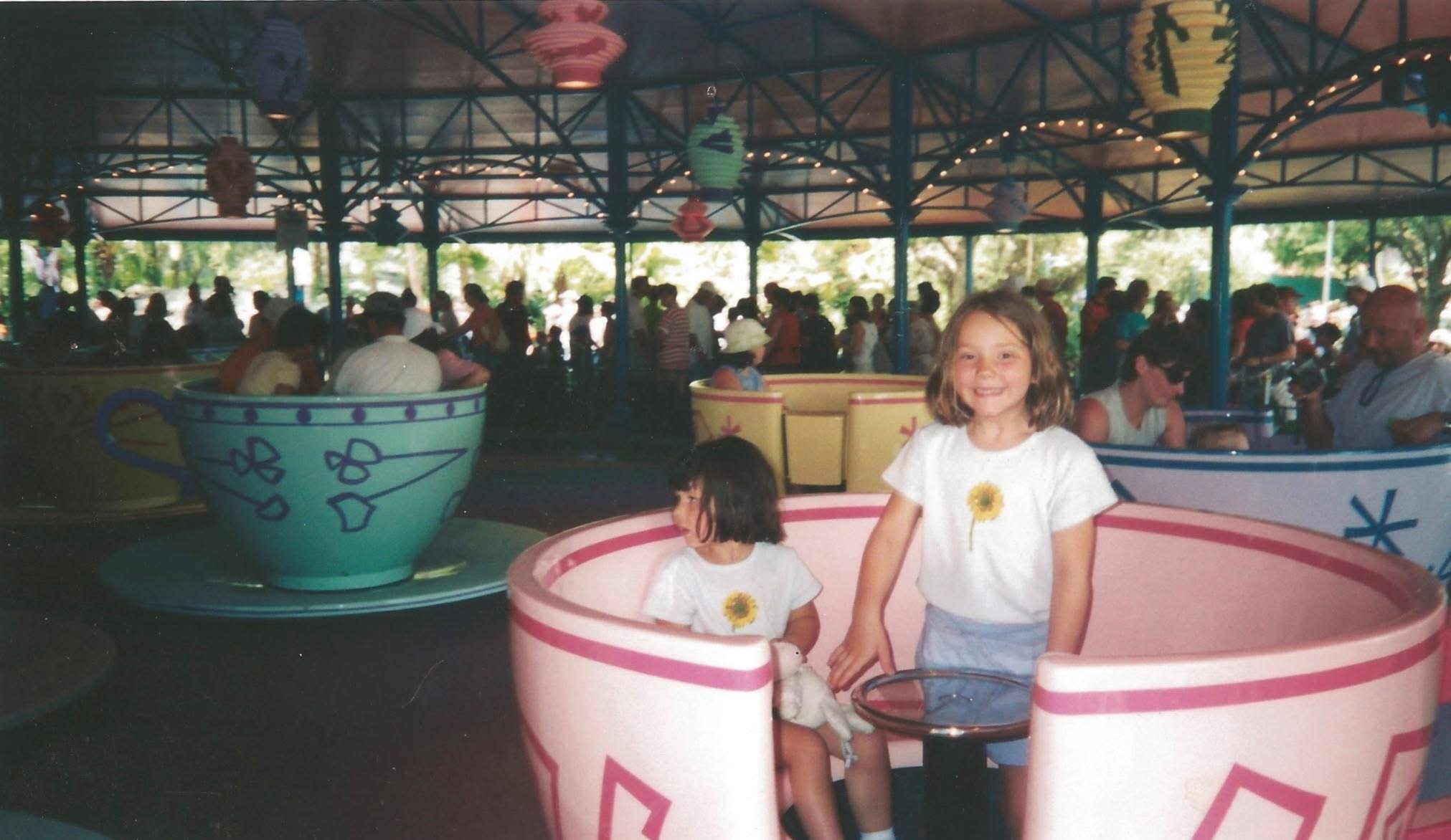 Two young girls in white t-shirts with yellow flower designs sitting in a pink teacup-shaped amusement park ride, with a girl smiling at the camera and a girl looking away. The background shows a crowded covered area with colorful lantern decorations hanging from the ceiling.