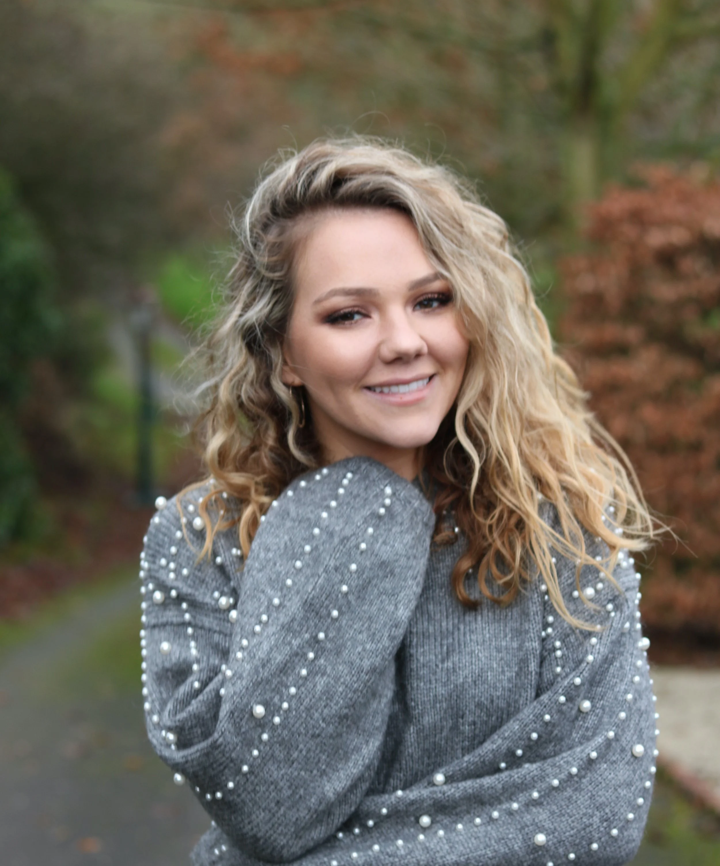 A smiling woman with curly blonde hair, wearing a gray sweater decorated with white pearls, outdoors in a natural setting with blurred trees in the background.