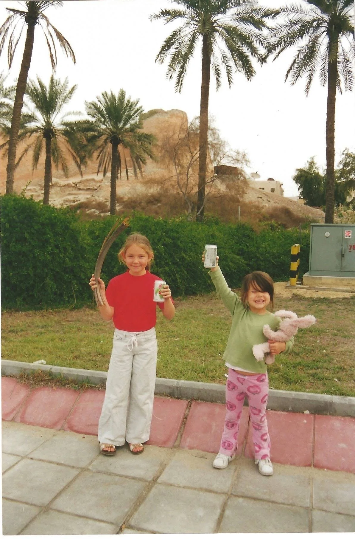 Two young girls standing outdoors on a sidewalk, holding drinks and a stuffed toy, with palm trees and dry landscape in the background.