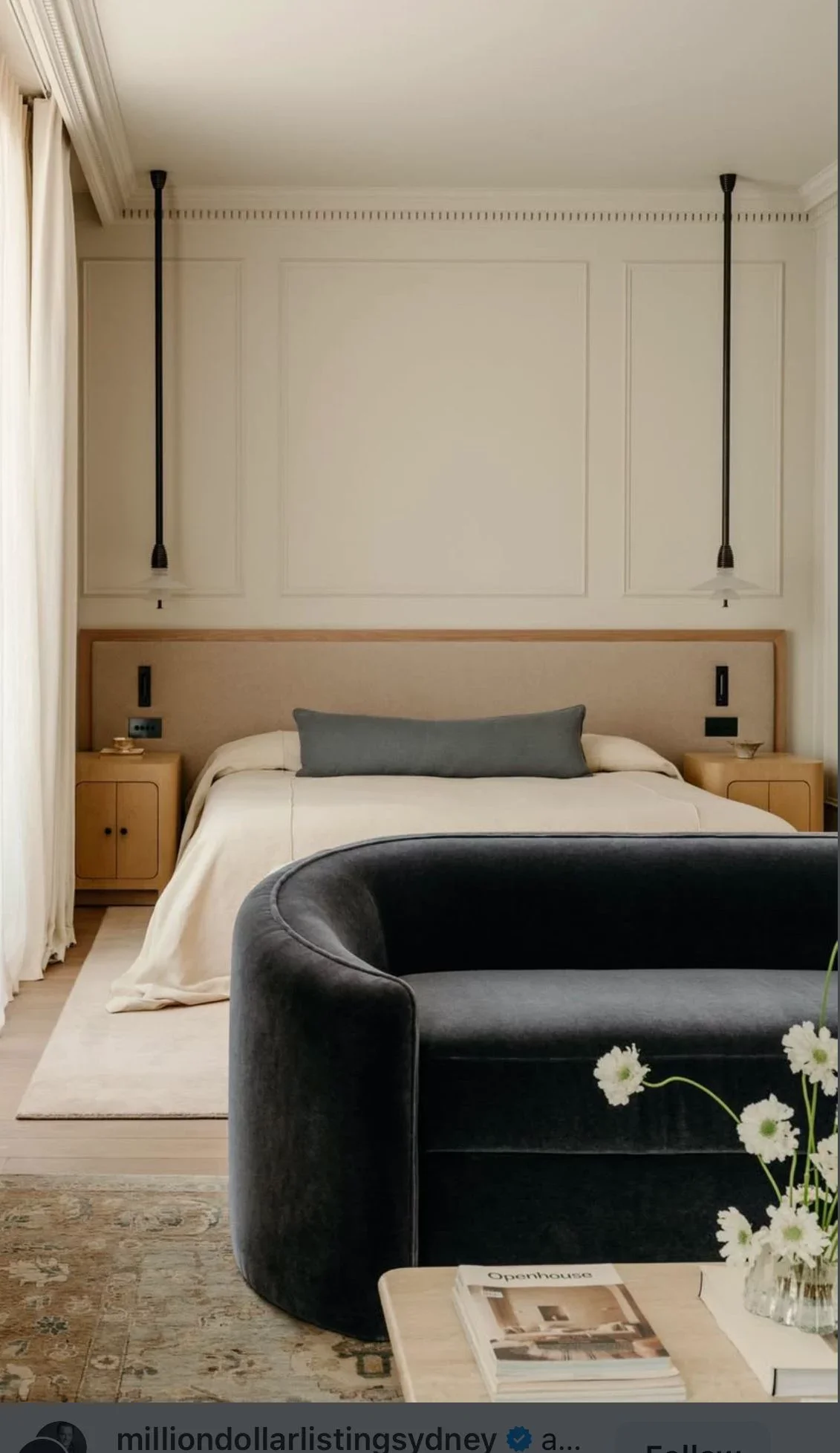 A modern bedroom with a bed, gray and white bedding, two bedside tables, a black armchair in the foreground, a vase with flowers, and hanging black pendant lights.