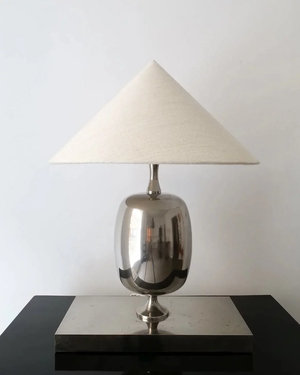 Silver table lamp with a white fabric shade, placed on a black and white table against a plain white wall.