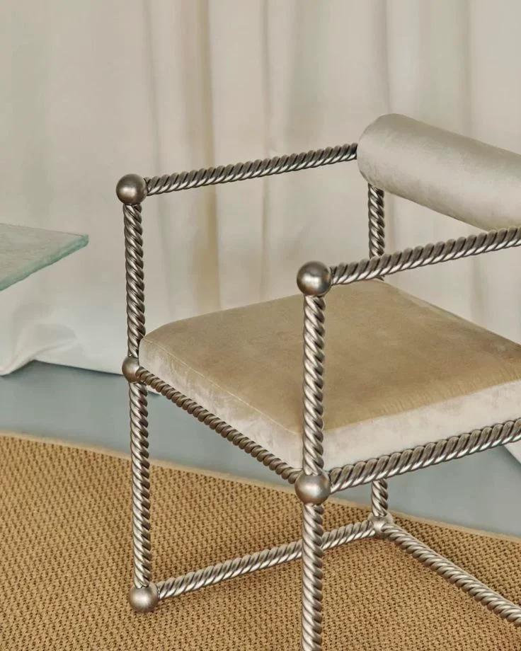 A modern fashionable chair with a metallic twisted rod frame and a beige velvet cushion, placed on a textured orange rug with a background of white curtains.