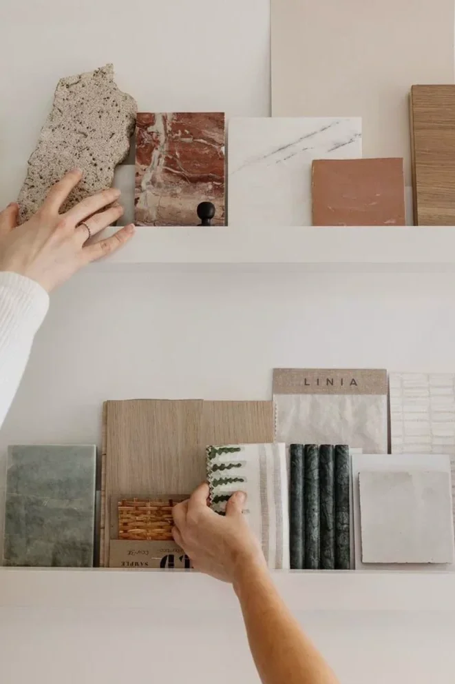 Two hands selecting tile samples from white display shelves. The samples include various textures, colors, and materials such as stone, marble, wood, and ceramic.