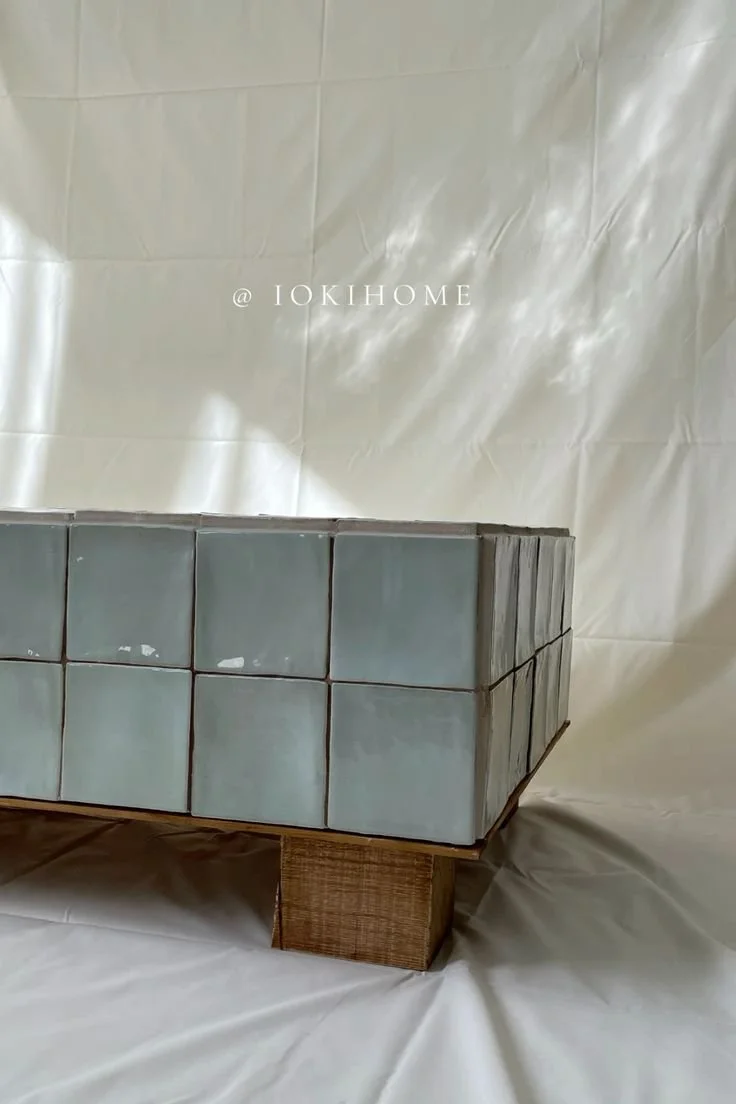 A large, rectangular surface made of light blue ceramic tiles, supported by a wooden block, with a crinkled white backdrop and the text '@ IOKIHOME' printed on it.