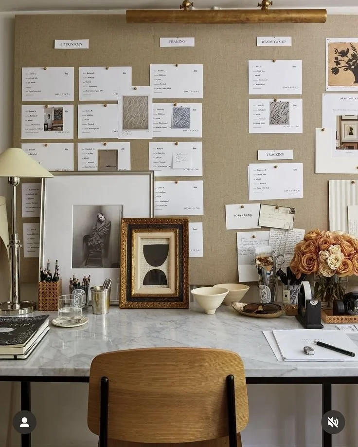 A workspace with a marble desk, framed art, a bouquet of roses, and a cork board wall with notes and photos organized in sections labeled 'In Progress', 'Framing', 'Ready to Ship', and 'Tracking'.