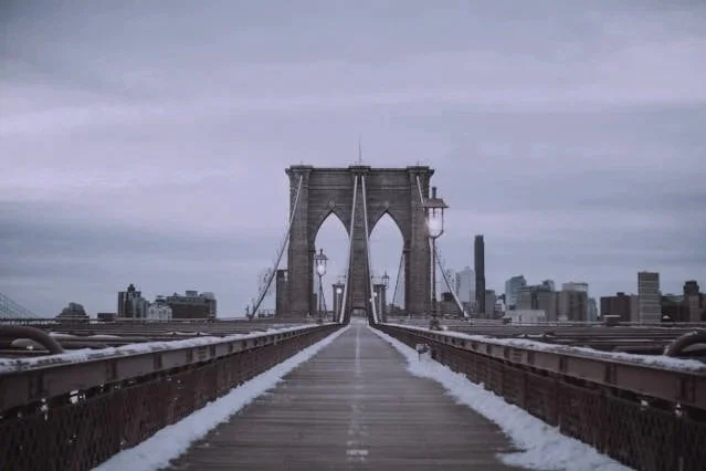 View of the Brooklyn Bridge with a wooden pedestrian walkway, city skyline in the background, and snow along the sides of the walkway under a cloudy sky.