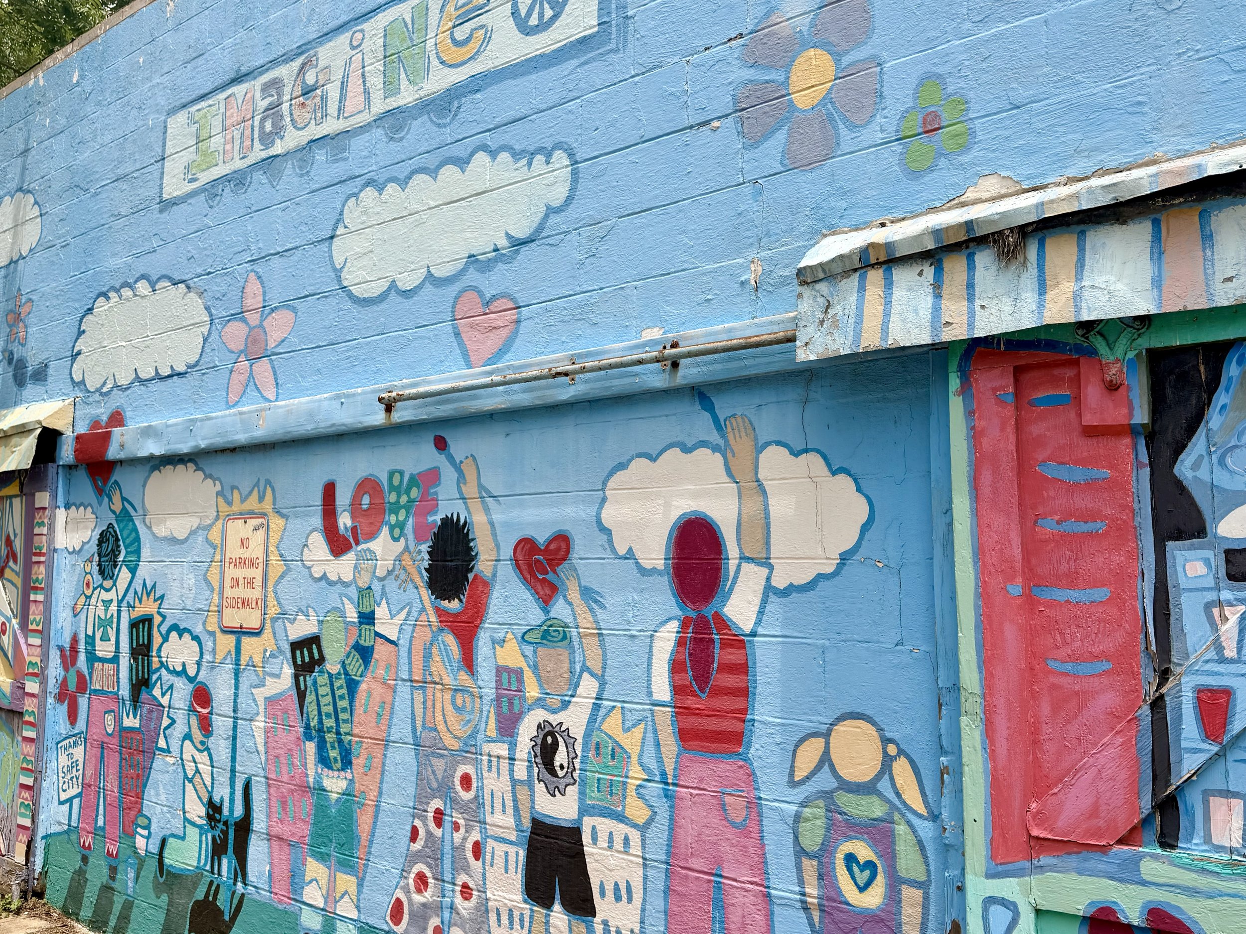 Colorful mural on a wall featuring clouds, flowers, hearts, buildings, and various illustrations, with the word 'IMAGINE' at the top.