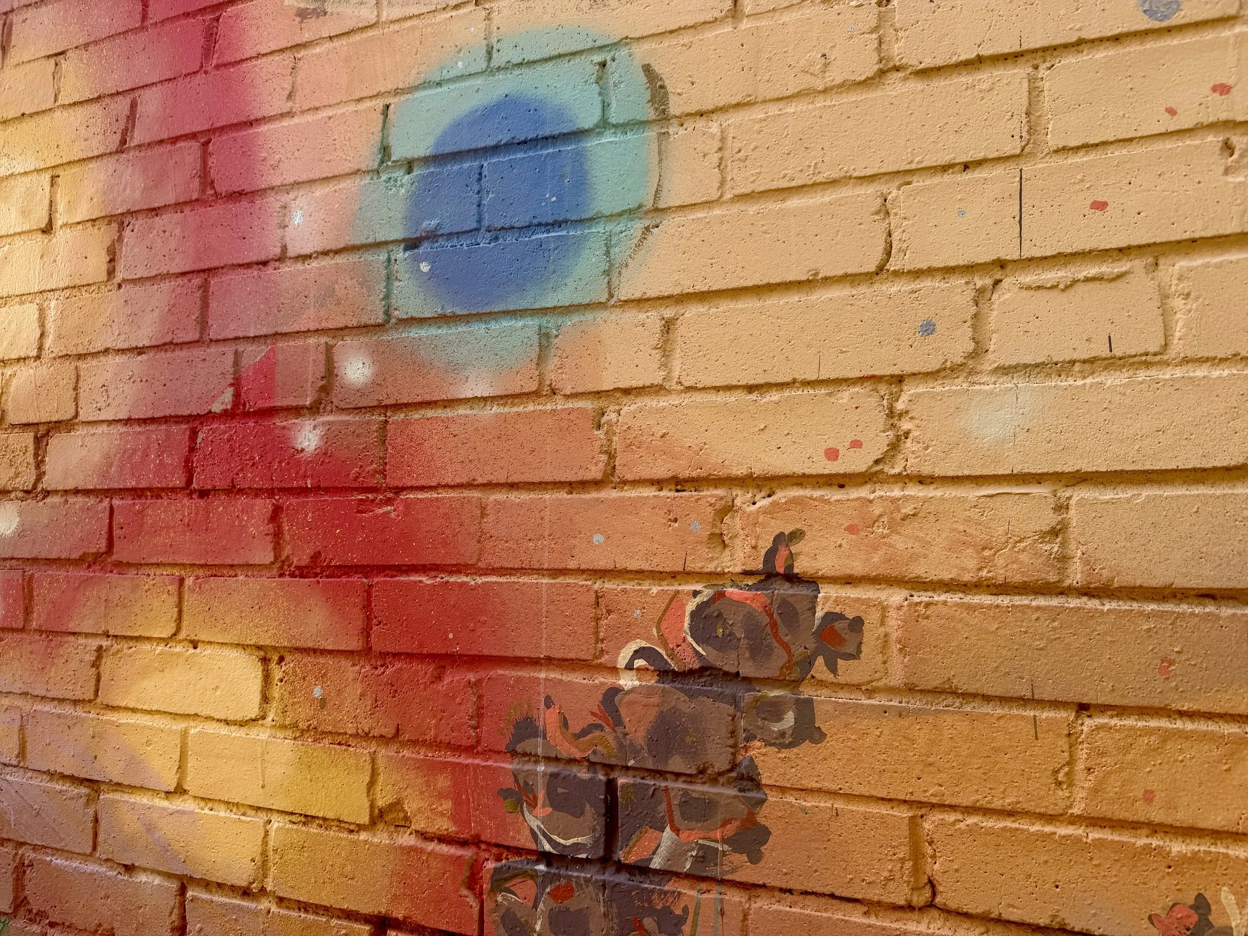 A brick wall painted in orange with a colorful graffiti art of a person's face with dark hair and glasses, with rainbow-colored circles and splatters surrounding it.