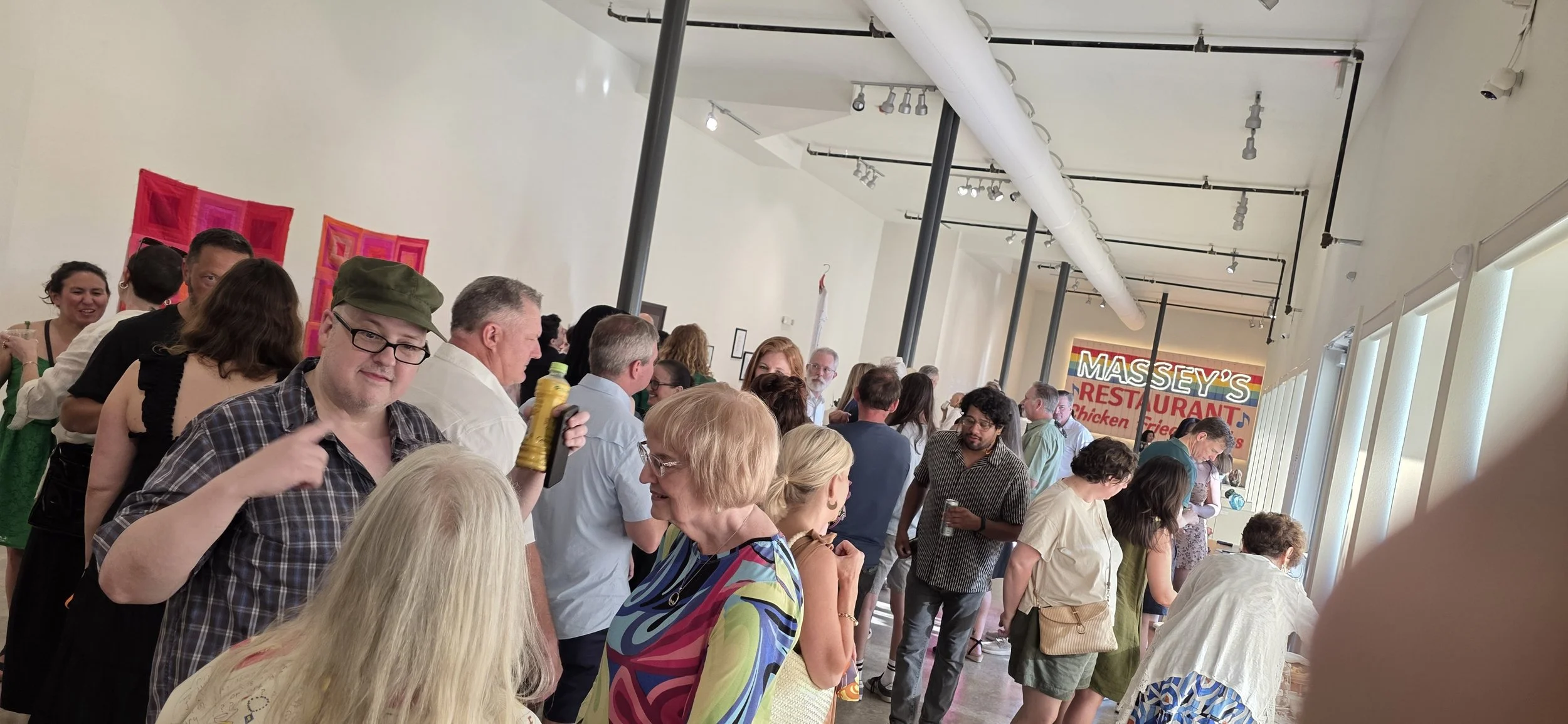A crowded indoor gathering with people socializing and chatting, some holding drinks. In the background, there is a sign for Massey's restaurant. The room has white walls, large windows on the right, and colorful artwork on the wall.