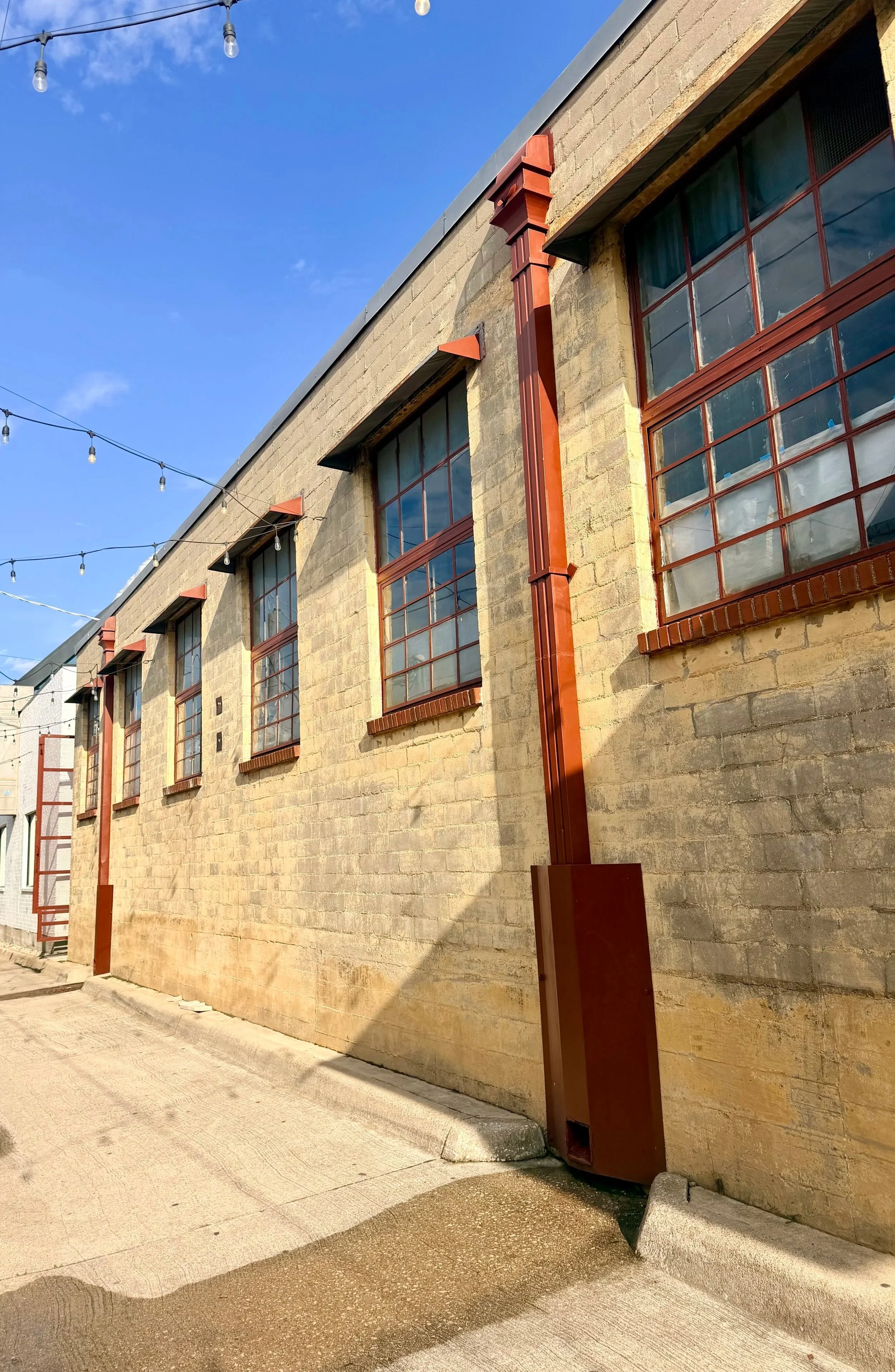 Exterior view of a brick building with multiple windows and reddish-brown drain pipes under a bright blue sky with string lights overhead.