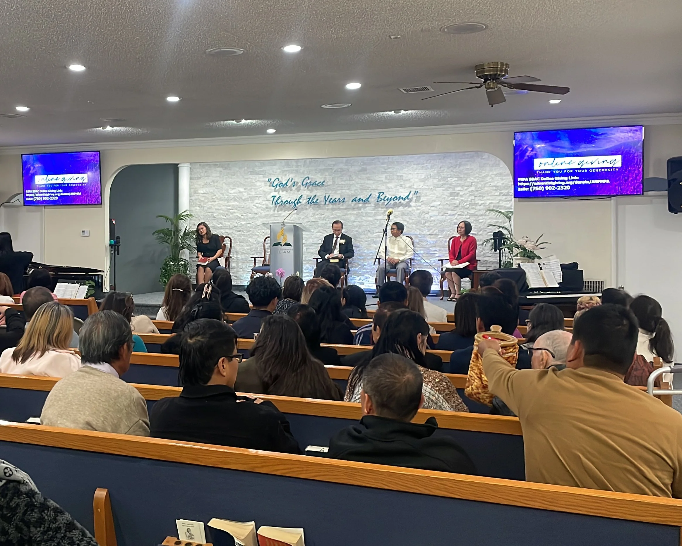 A church service or conference taking place inside a church or conference hall, with several people seated. Four individuals are seated on stage, with two women and two men. The stage features a white brick backdrop with the words 'God's Grace Through the Years and Beyond' written on it, and two large screens displaying the same message and contact information. The audience is seated in wooden pews, and there are potted plants on stage. Overhead lighting and a ceiling fan are also visible.