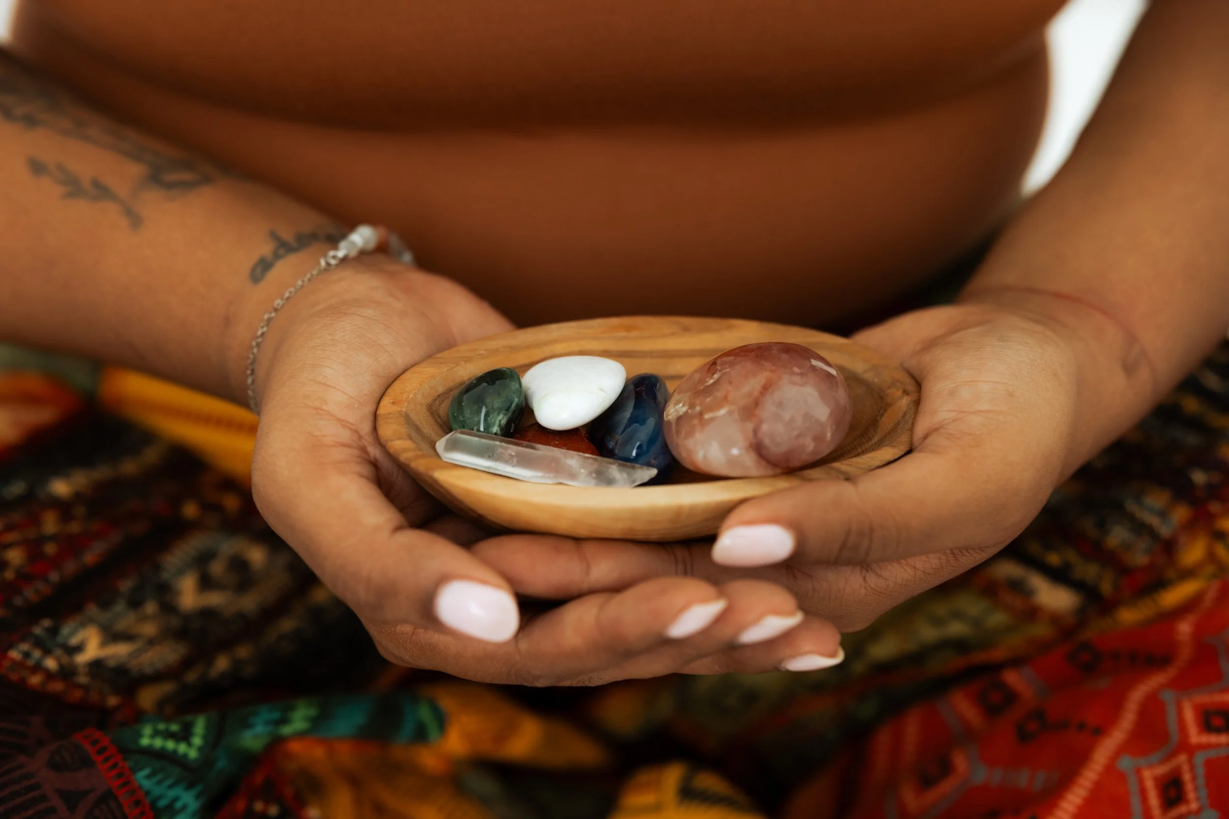 A person with tattoos on their arm is holding a small wooden dish containing polished stones and crystals, including green, white, blue, and reddish stones. The person has a silver bracelet on their wrist and white nail polish, and they are wearing colorful clothing with a patterned fabric in the background.