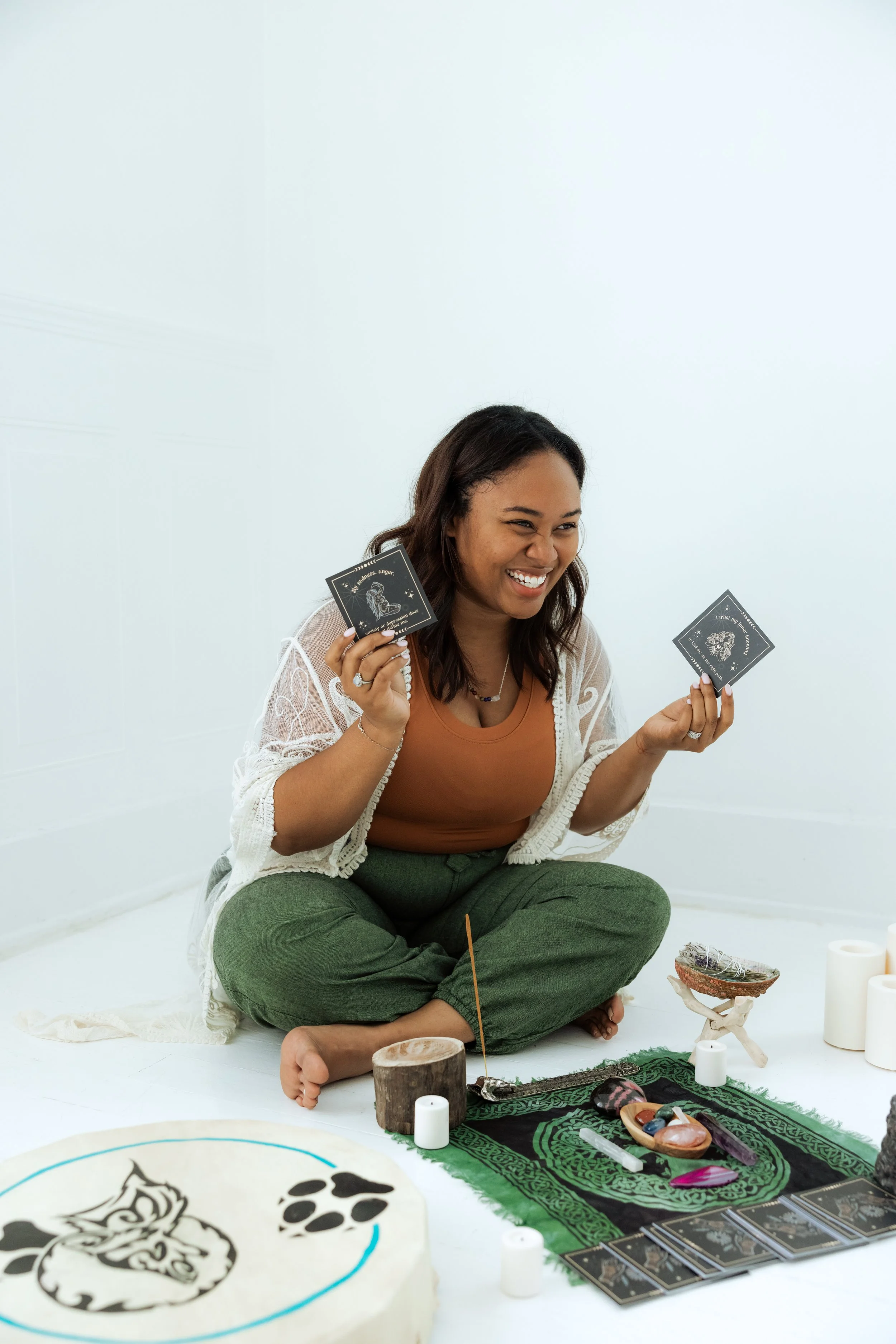 A woman sitting on the floor with her legs crossed, smiling and holding tarot cards. She is surrounded by candles, stones, and other spiritual items, with a white background.