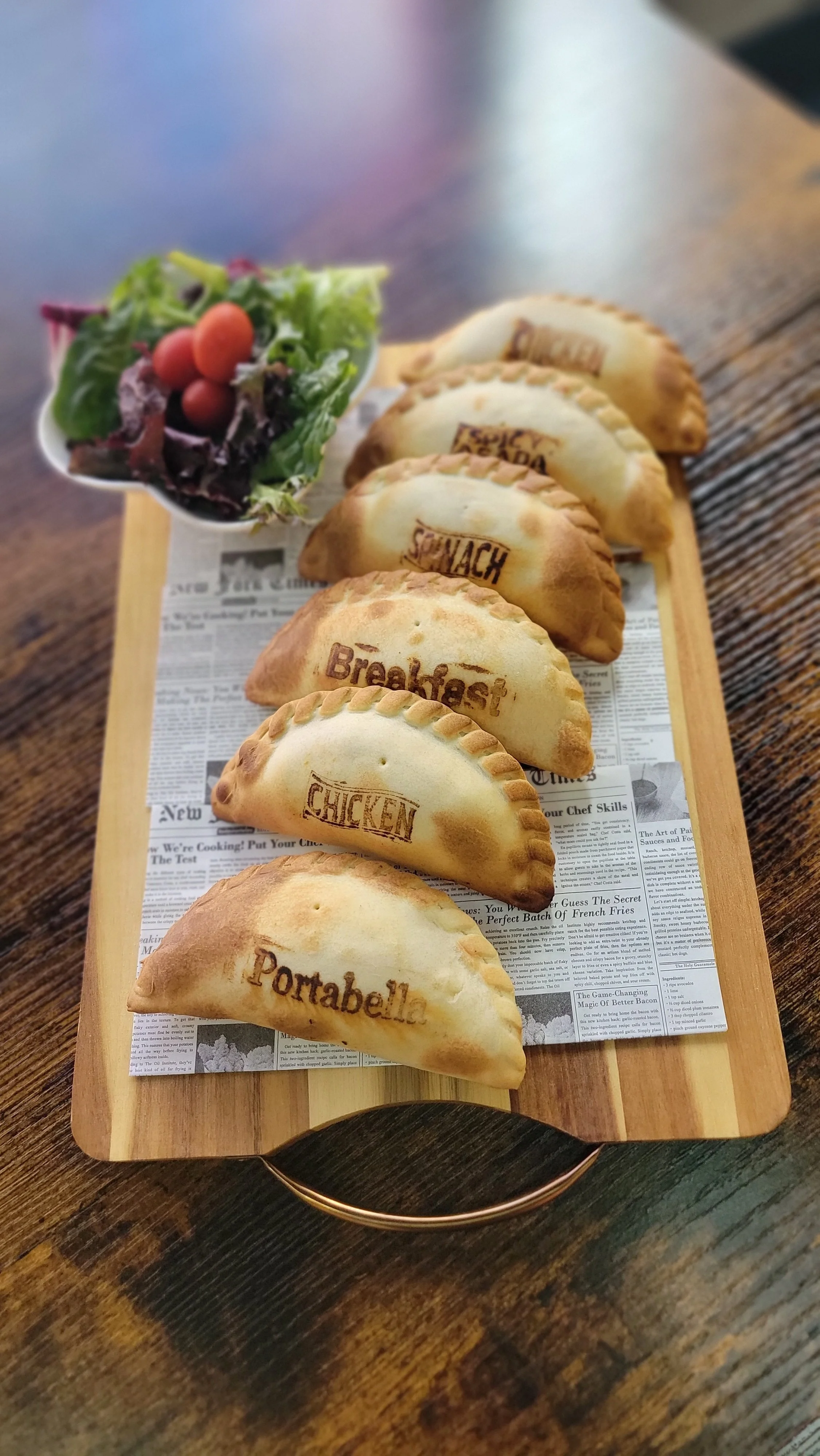 A wooden serving board with fried empanadas labeled 'Pottabella', 'Chicken', 'Breakfast', and 'Savory', arranged in a row, on a dark wooden table with a side salad of lettuce, cherry tomatoes, and mixed greens