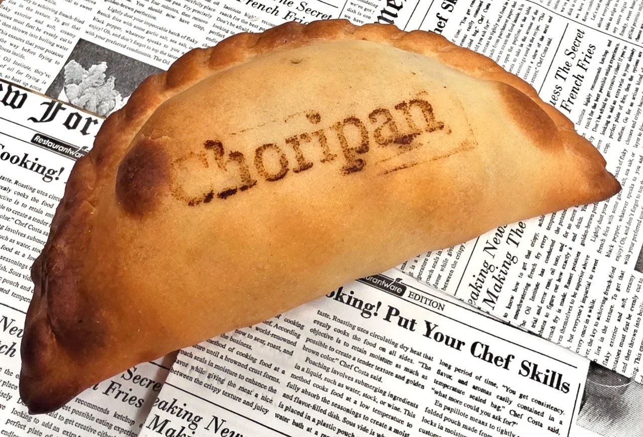 A golden-brown fried empanada with the word 'Chopin' burned onto its surface, placed on a bed of black-and-white newspaper pages.