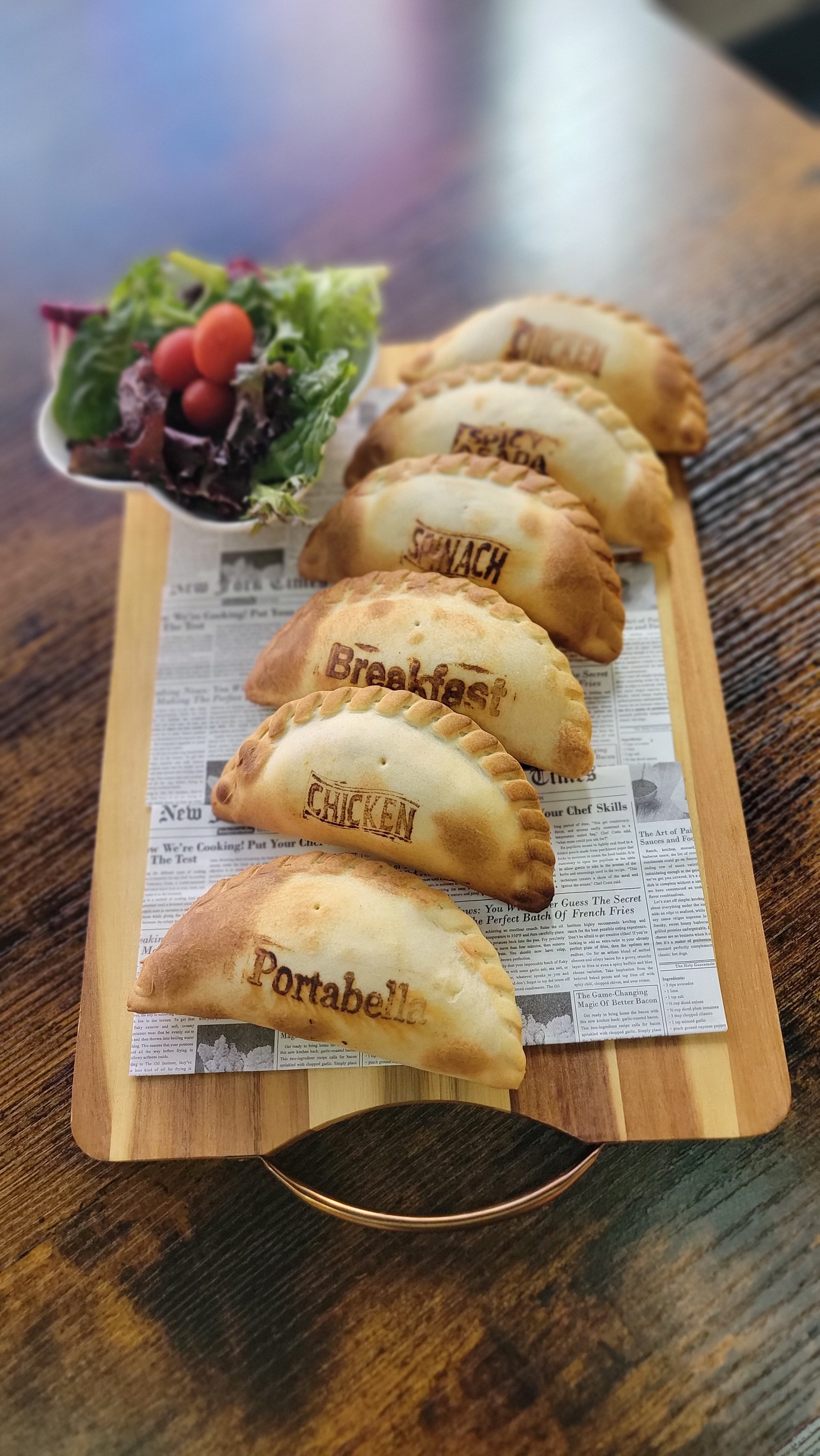A wooden tray with baked empanadas labeled as Chicken, Breakfast, Portabella, and Potabella, and a small bowl of mixed greens salad with cherry tomatoes.