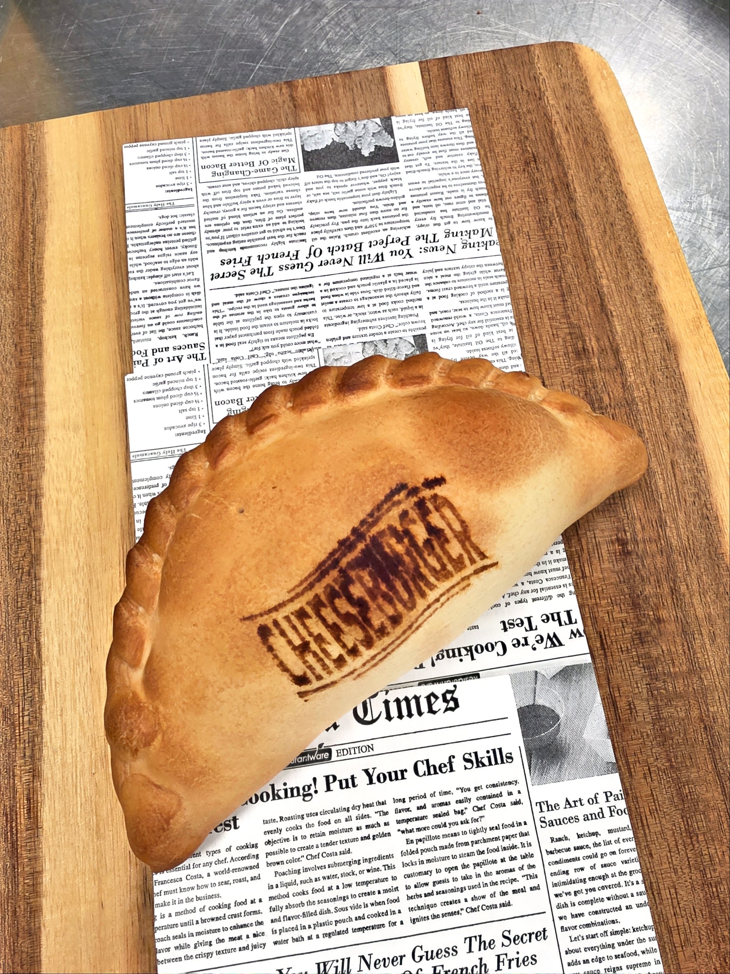 A golden-brown hand pie with a crimped edge and a stamped logo on top, placed on a wooden cutting board with a background of a newspaper.