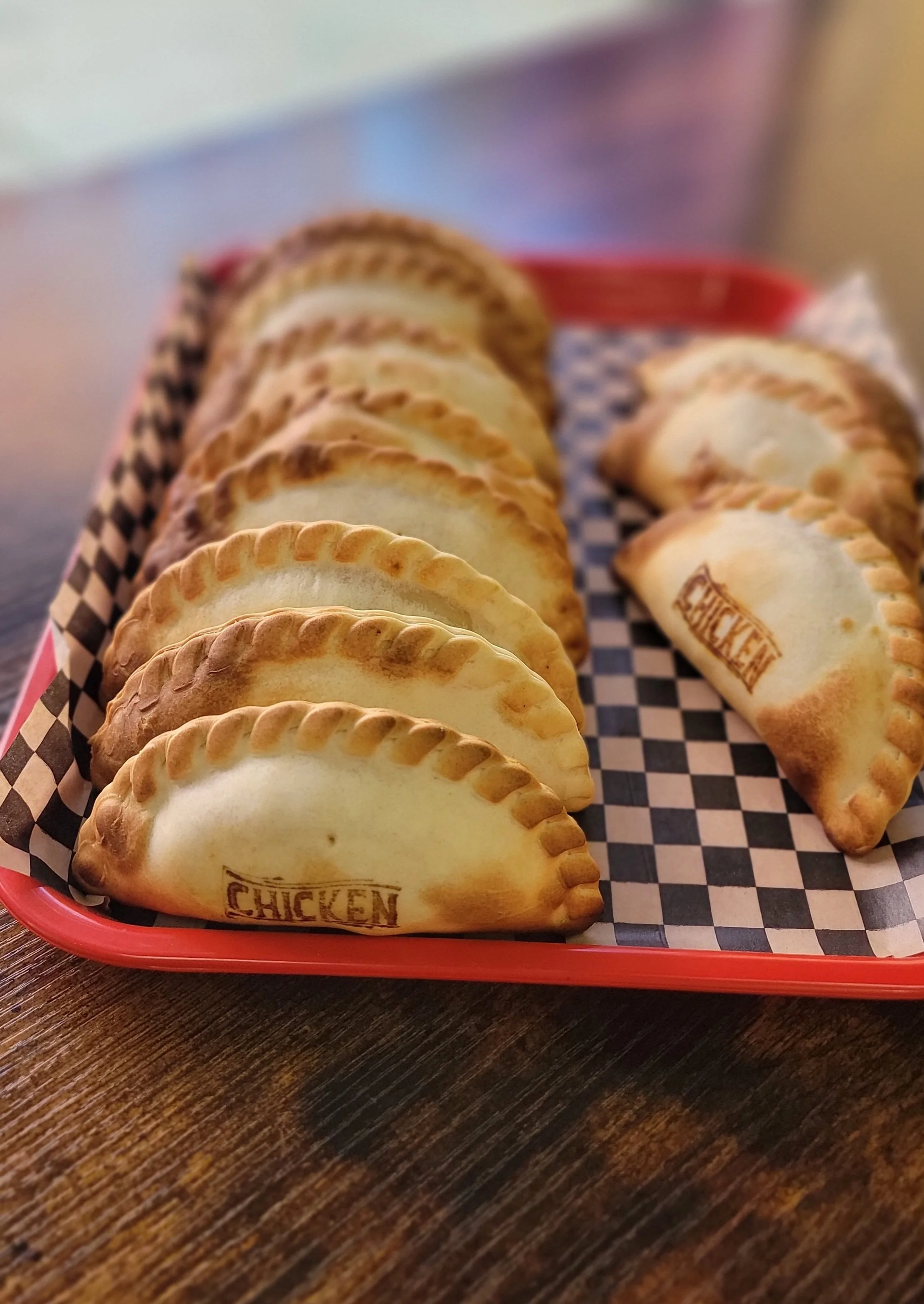 Close-up of a red basket with checkered paper lining, holding several baked empanadas with golden-brown crusts and the word 'CHICKEN' stamped on one of them, placed on a wooden surface.