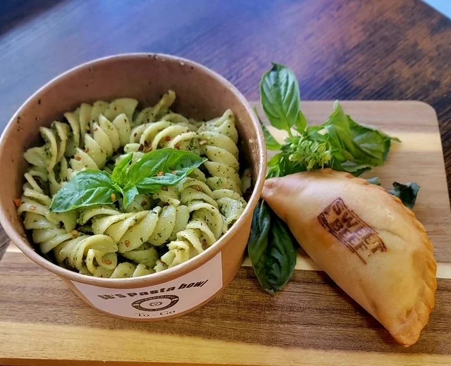 Bowl of pesto pasta garnished with basil leaves, placed on a wooden serving board with fresh basil and a half-moon-shaped fried snack on the side.