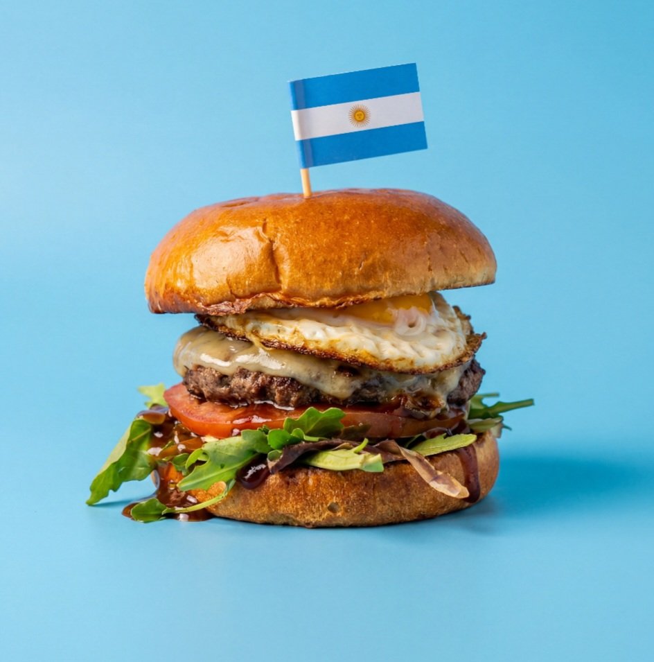 A burger with a fried egg, tomato, lettuce, and cheese, topped with an Argentinian flag on a skewer