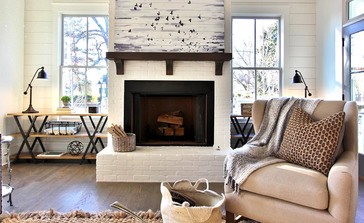 Living room with white brick fireplace, wooden mantel, black firebox, beige sofa with patterned pillow, throw blanket, side shelves with lamps, windows showing trees and outdoor scene.