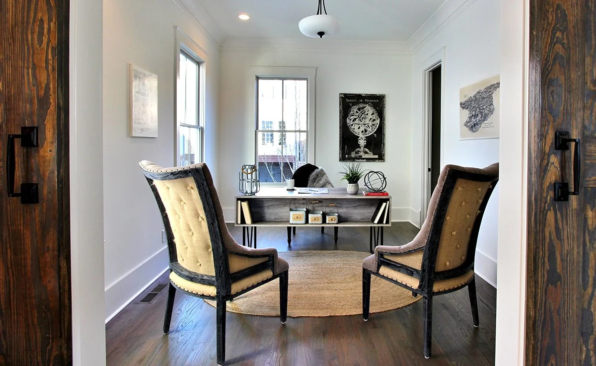 Living room with two vintage armchairs, a wooden desk, artwork on the walls, a window, and a rug on wooden flooring.
