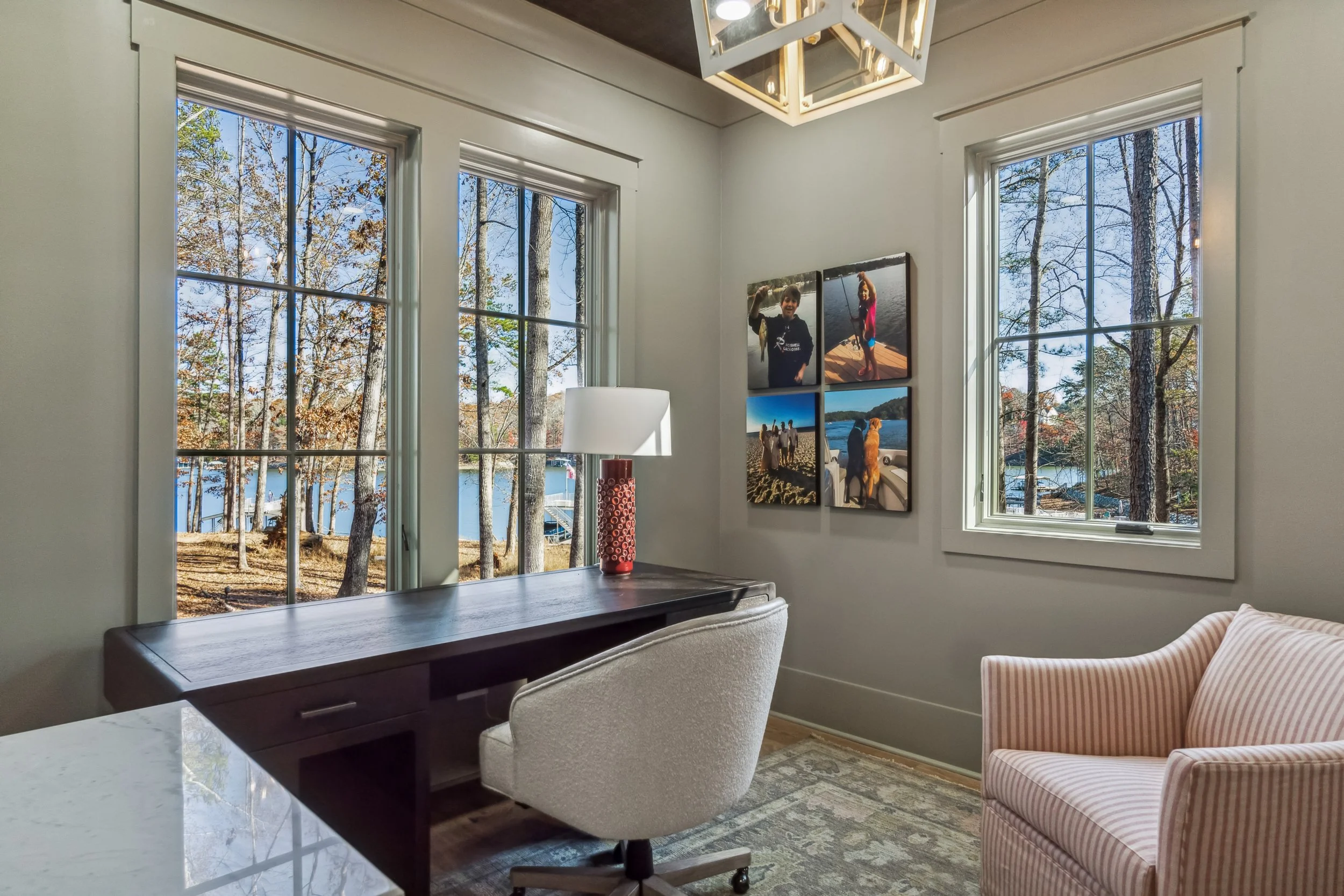 A room with three large windows showing a view of trees and a lake outside. Inside, there is a wooden desk with a white desk chair, a table lamp with a red base, a beige upholstered armchair, and a pink-striped armchair. A framed photo collage hangs 