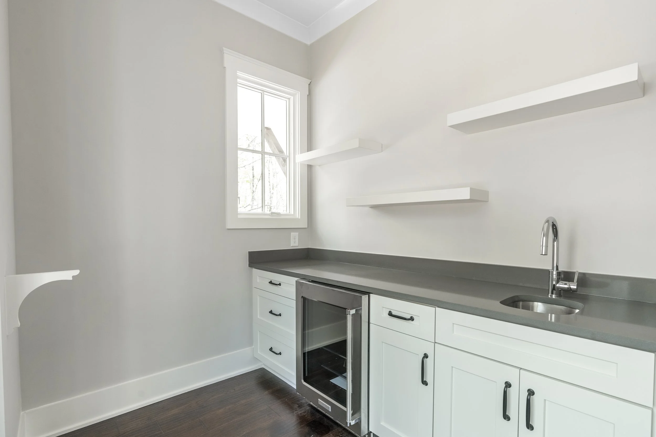 A modern kitchen with white cabinets, a gray countertop, a small wine refrigerator, a stainless steel sink, a chrome faucet, and two white floating shelves on a white wall by a window with sunlight.