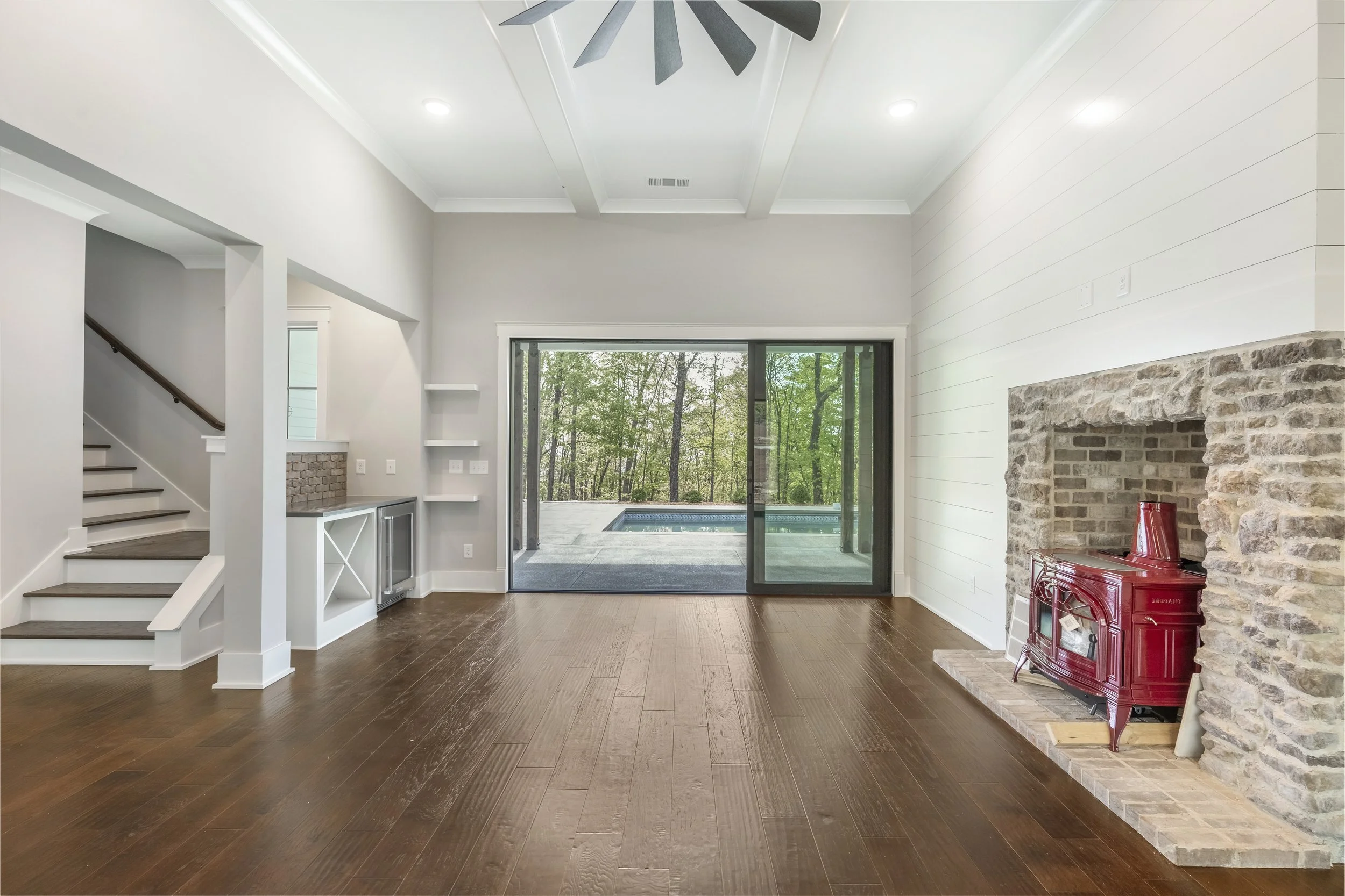 Bright living room with hardwood floors, a stone fireplace with a red wood stove, large windowed doors leading outside to a deck with a pool, and stairs leading upstairs.