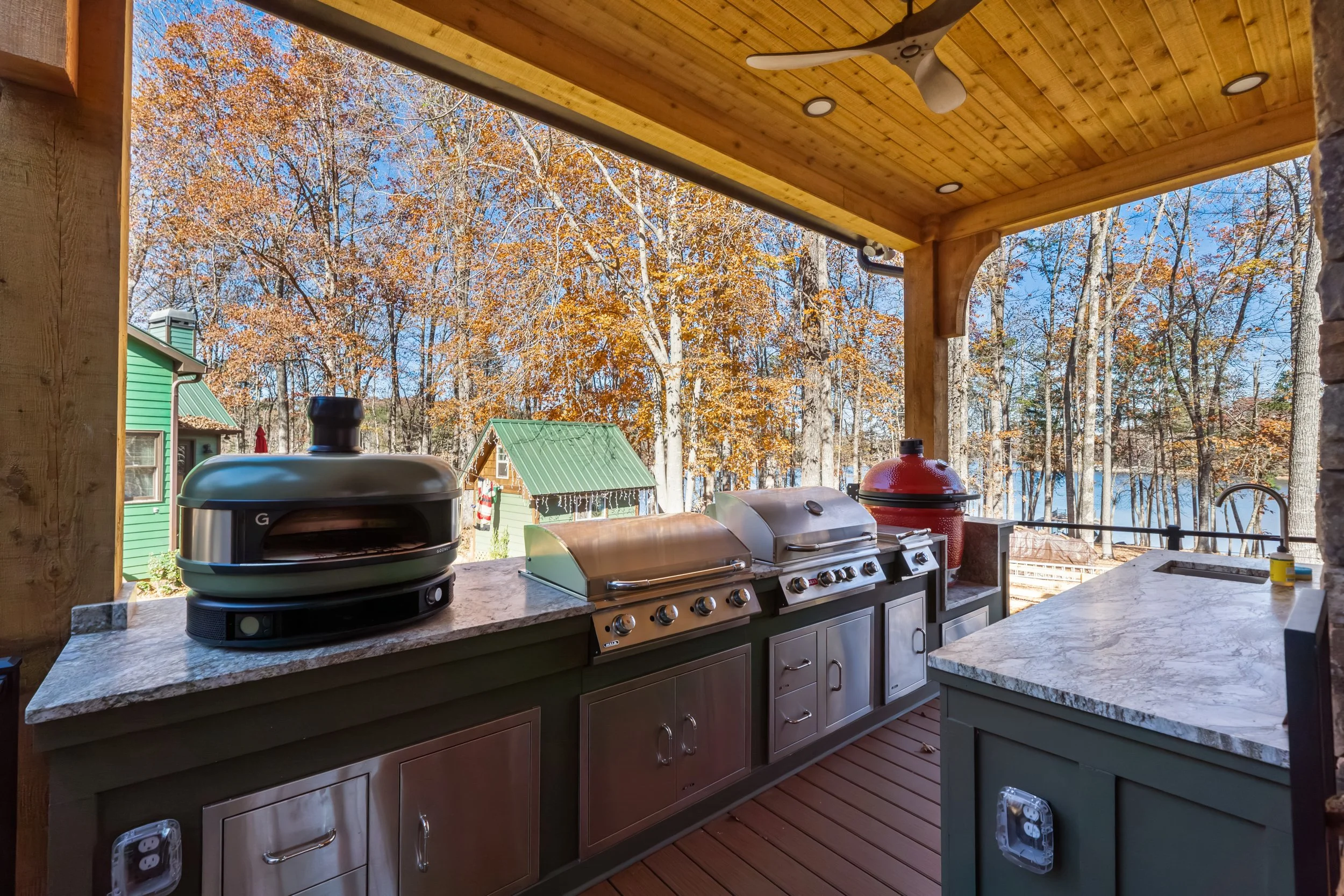 Outdoor kitchen area with stainless steel grill, smoker, and countertop with a sink. Wooden ceiling with recessed lighting and a ceiling fan. View of trees with fall foliage and small green shed in the background.