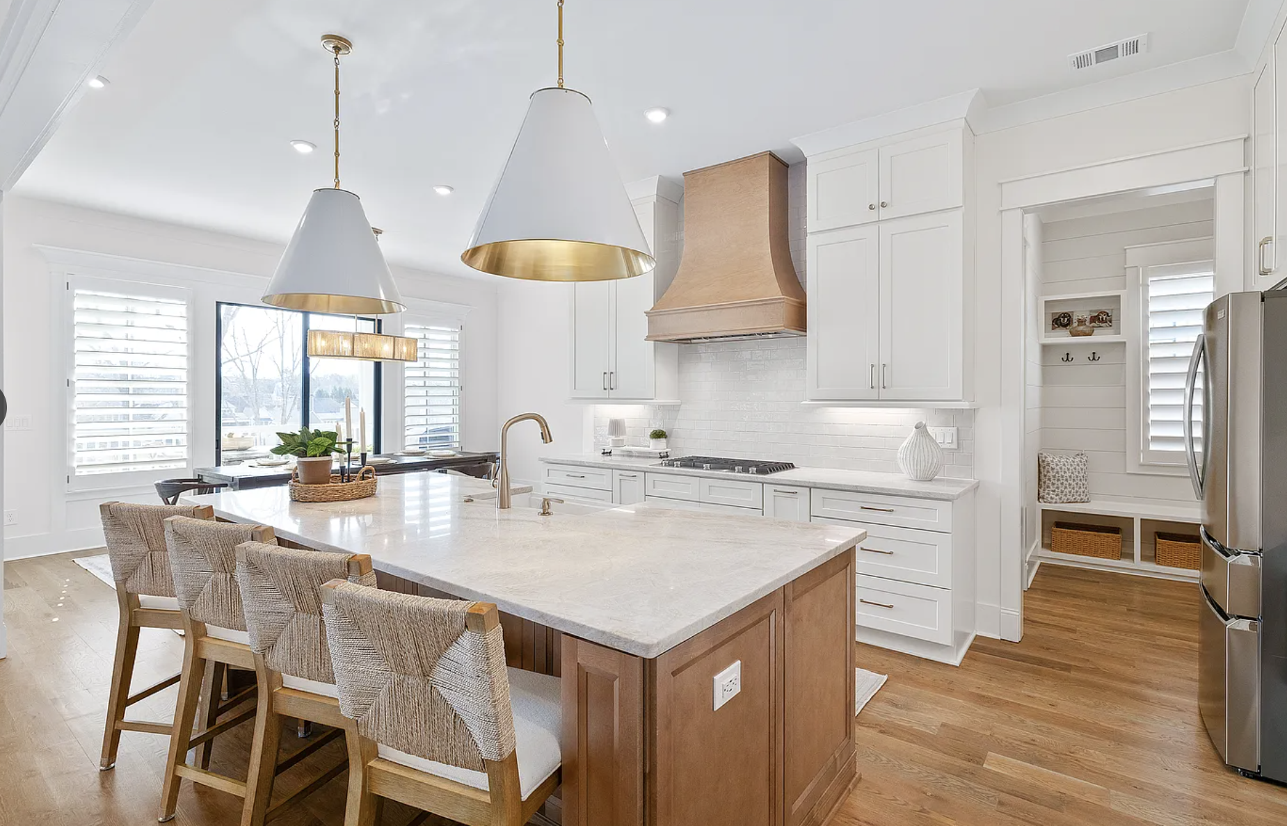 Modern white kitchen with a large island, four wicker chairs, pendant lighting, and a window with shutters, hardwood flooring, and decorative accessories.