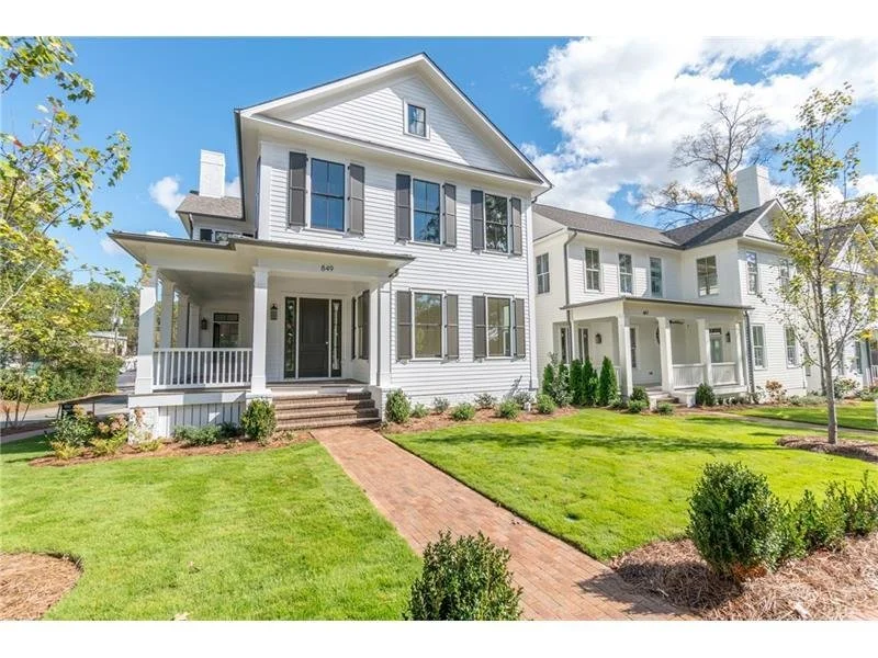 A white two-story house with black shutters, black front door, and a front porch with stairs, surrounded by a well-maintained lawn and small bushes, under a partly cloudy sky.