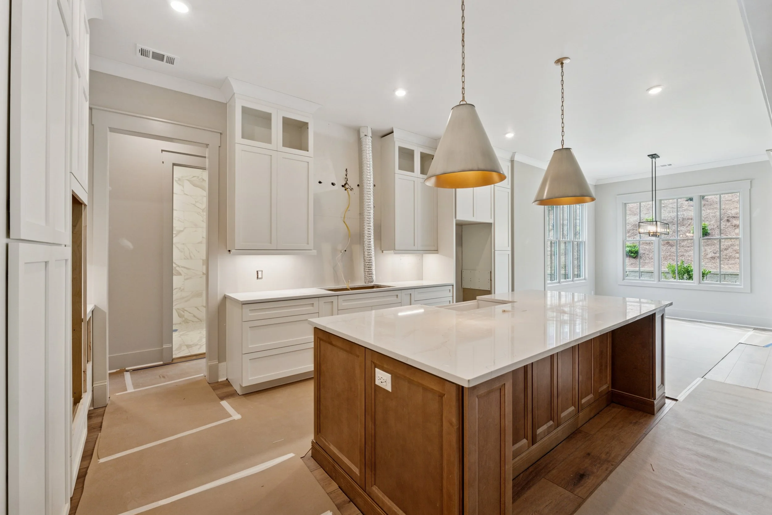 Interior of a kitchen under construction with white cabinets, a wooden island, and large windows, with some finishing touches ongoing.