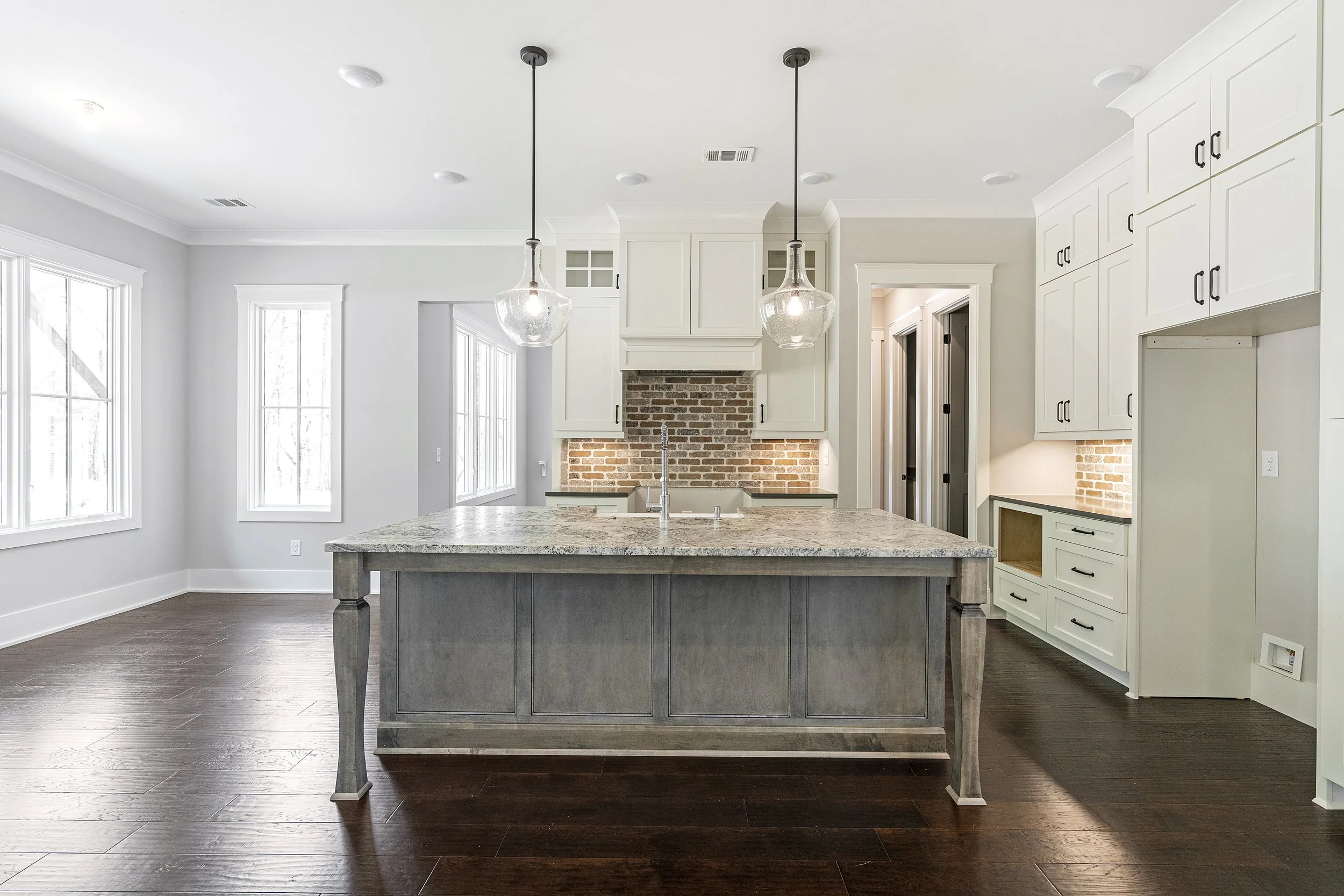 Modern kitchen with white cabinetry, a brick backsplash, and a large kitchen island with a granite countertop. Two pendant lights hang above the island. Large windows let in natural light, and dark hardwood floors complete the space.