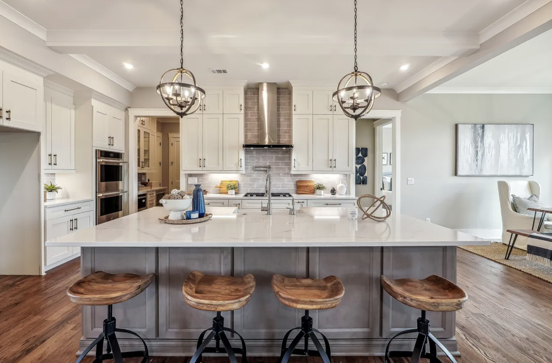 Modern kitchen with white cabinets, stainless steel appliances, and a large island with a marble countertop and four wooden stools. Two decorative chandeliers hang from the ceiling. There's a small seating area with a white chair and a large abstract