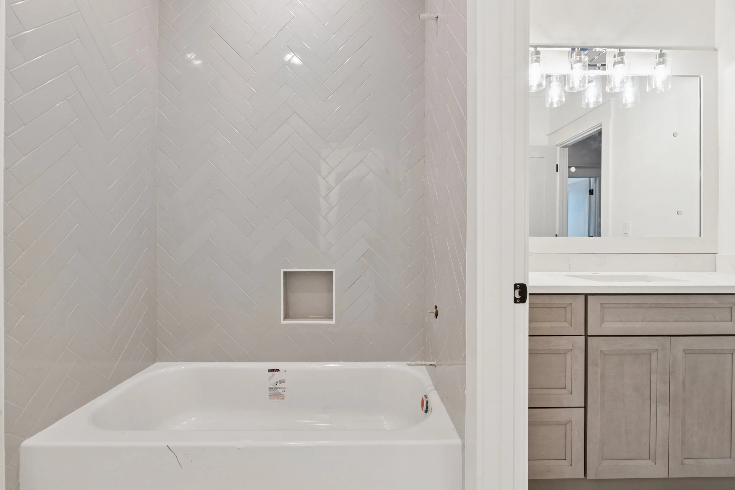 Empty bathroom with a bathtub, a wall with light-colored tiles arranged in a herringbone pattern, and a mirror above a vanity with wooden cabinets.