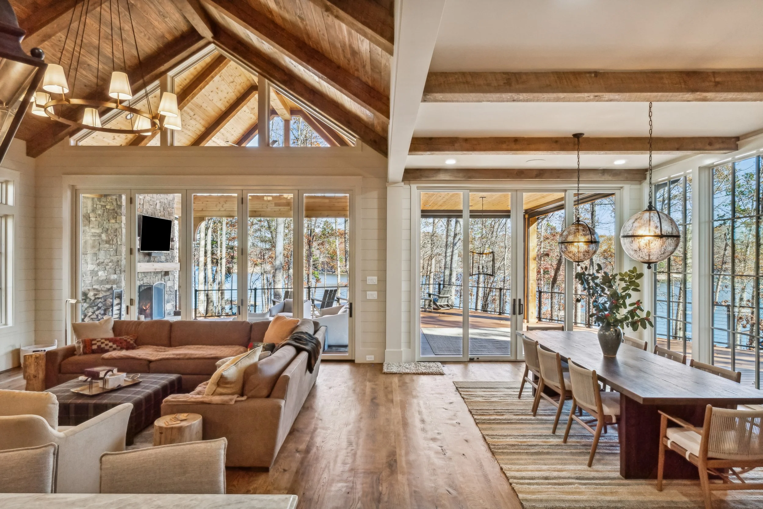 Interior view of a spacious living and dining area with large windows and glass doors, wooden beams, a stone fireplace outside, a sectional sofa, and a dining table with chairs. The room is filled with natural light.