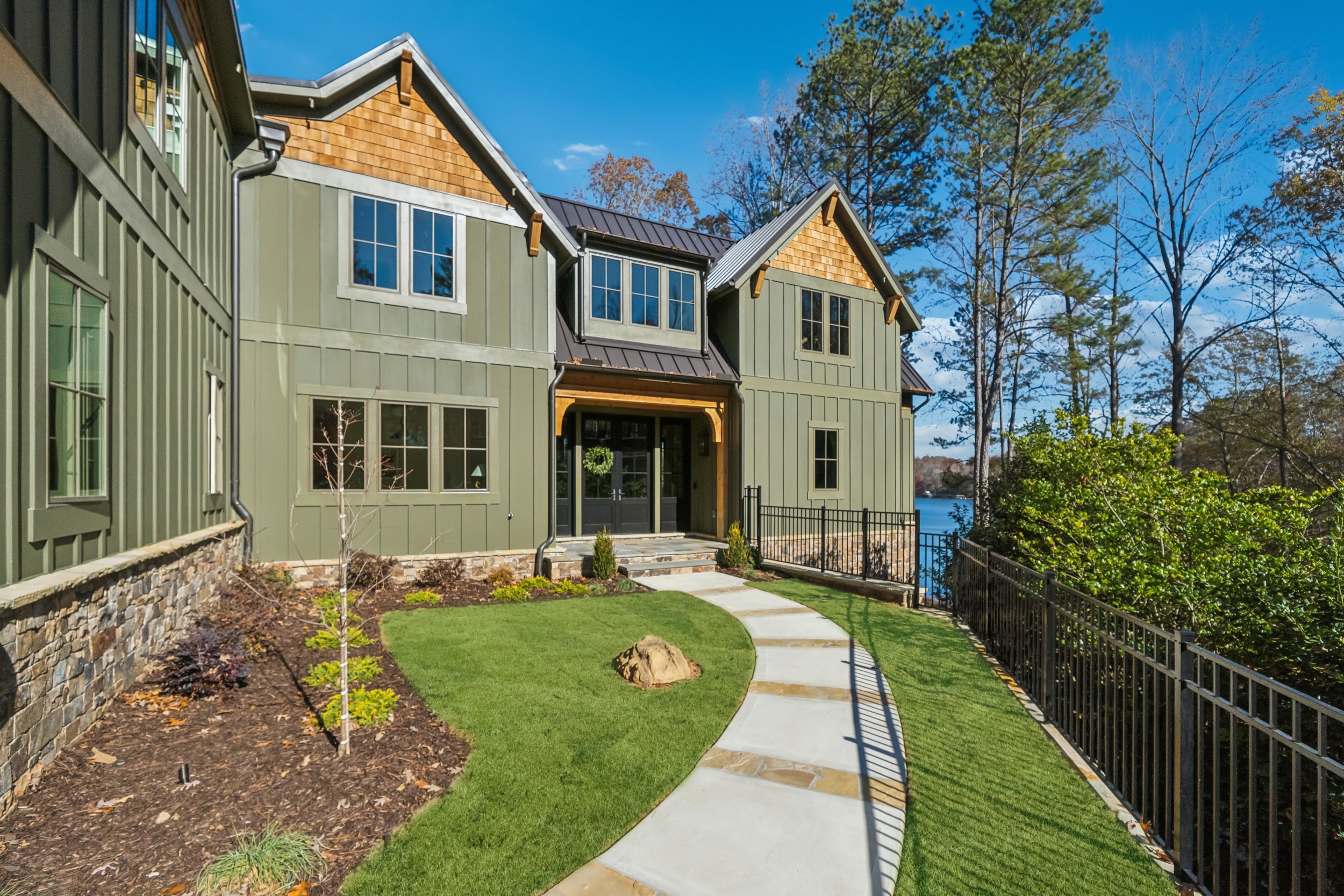 A residential house with green siding, divided into sections with gabled roofs, large windows, and a front porch. The house is near a body of water and surrounded by trees, with a landscaped yard featuring a stone-bordered flower bed and a curved concrete walkway.