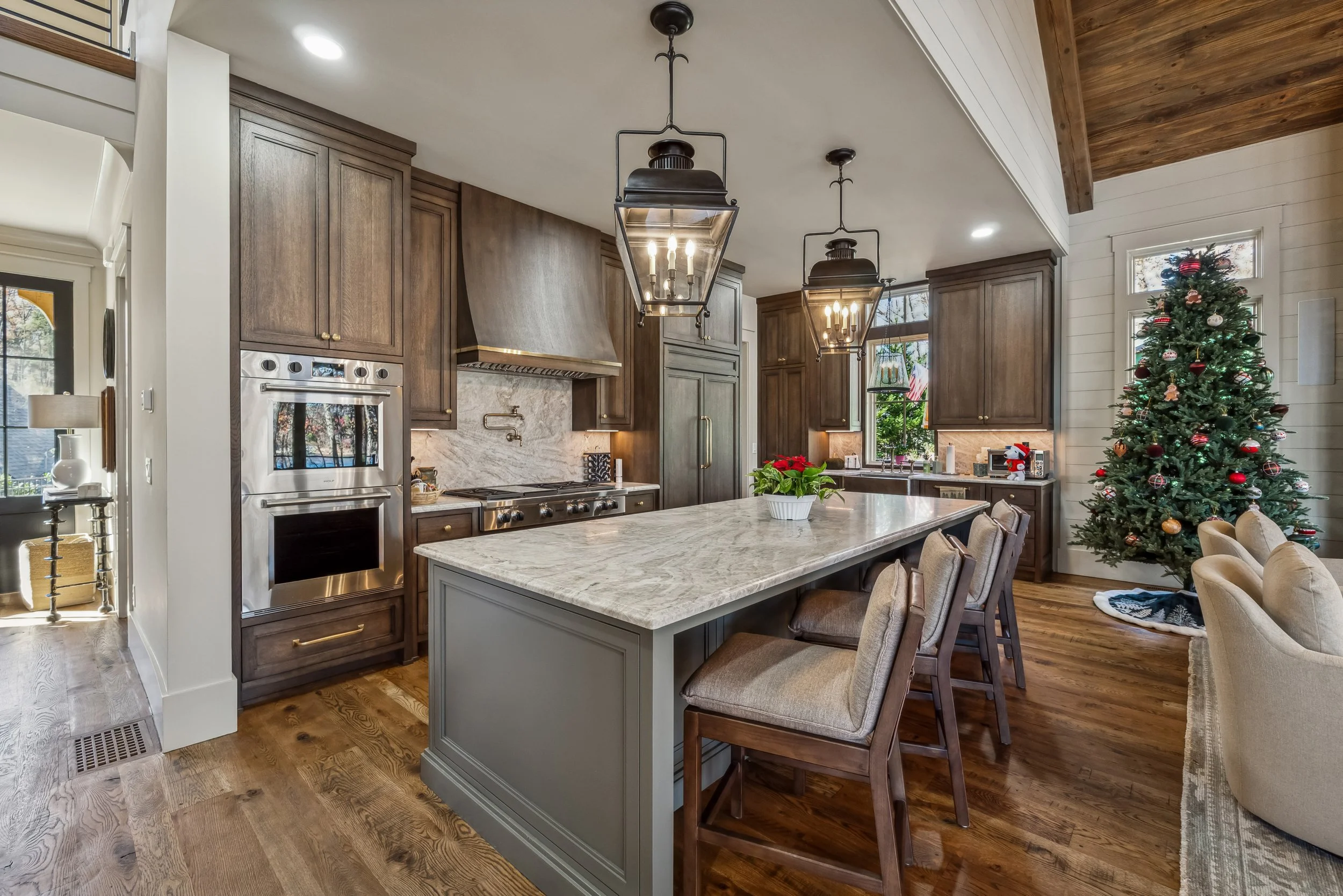 Open-concept kitchen with dark wood cabinets, a marble island, and two pendant lights hanging over the kitchen island. Decor includes a Christmas tree with ornaments, poinsettia plant on the island, and natural light from windows.