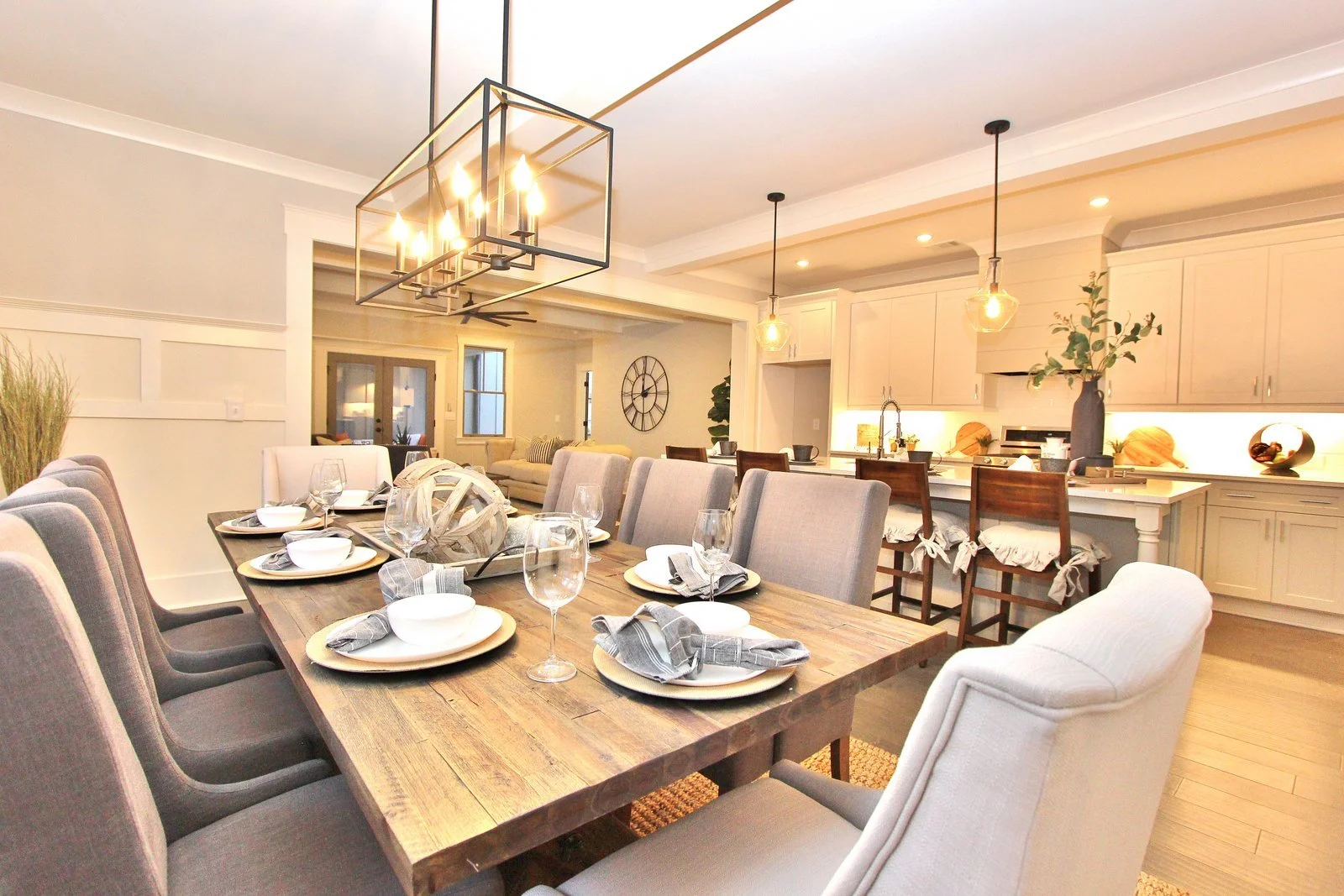 Open-concept dining and kitchen area with a large wooden table set with white dishes, glasses, and napkins, surrounded by upholstered chairs, and a modern chandelier overhead. In the background, a kitchen with white cabinets, a breakfast bar with bar