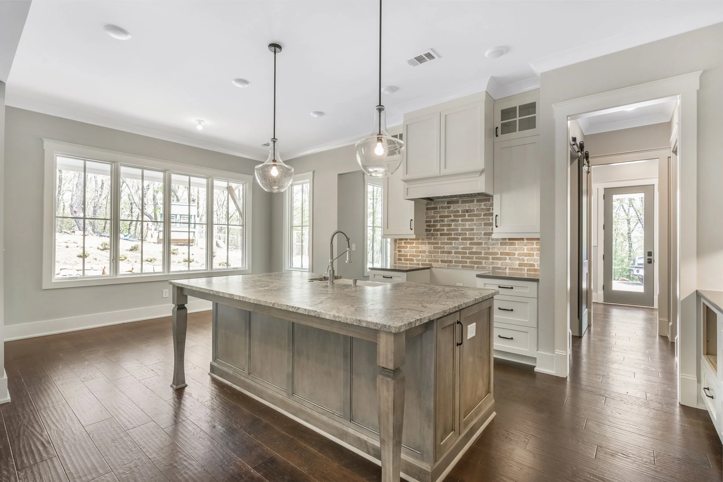 Modern kitchen with large windows, a kitchen island with a granite countertop, white cabinets, and a brick backsplash, illuminated by pendant lights, with hardwood floors and a view of trees outside.