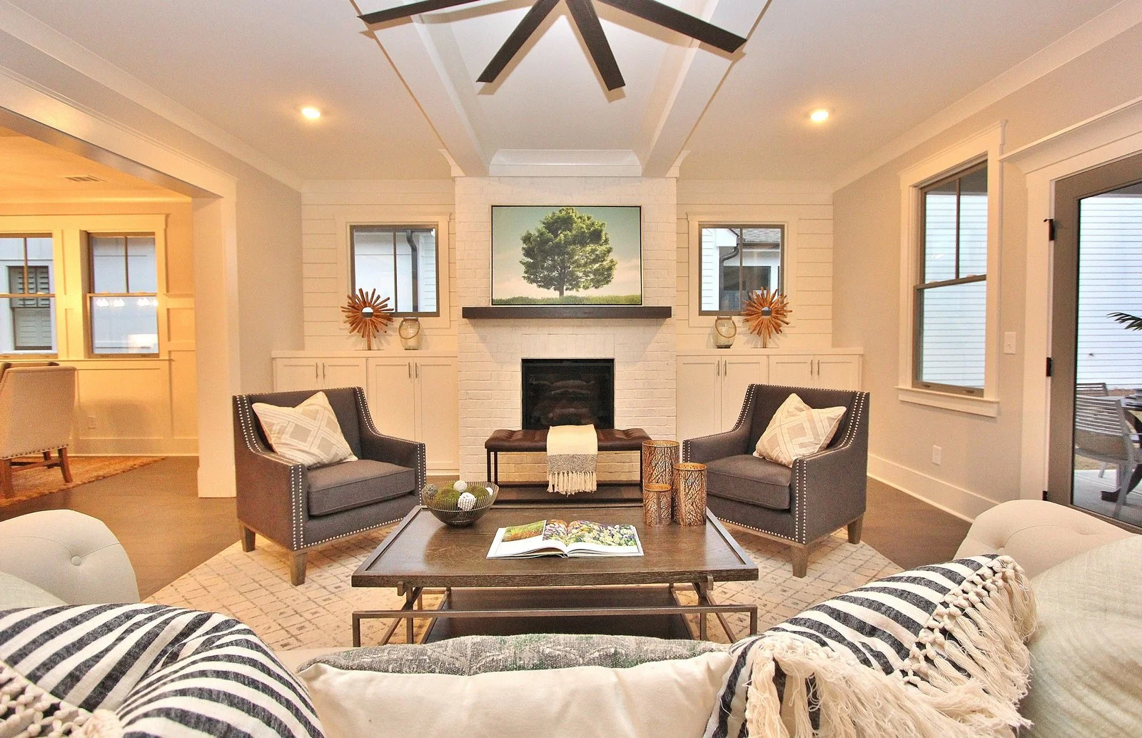 A modern living room with white brick fireplace, dark wood coffee table, two gray armchairs with pillows, and a wall-mounted TV above the fireplace showing a tree. There is a white sofa with a striped throw in the foreground. Large windows and a slid